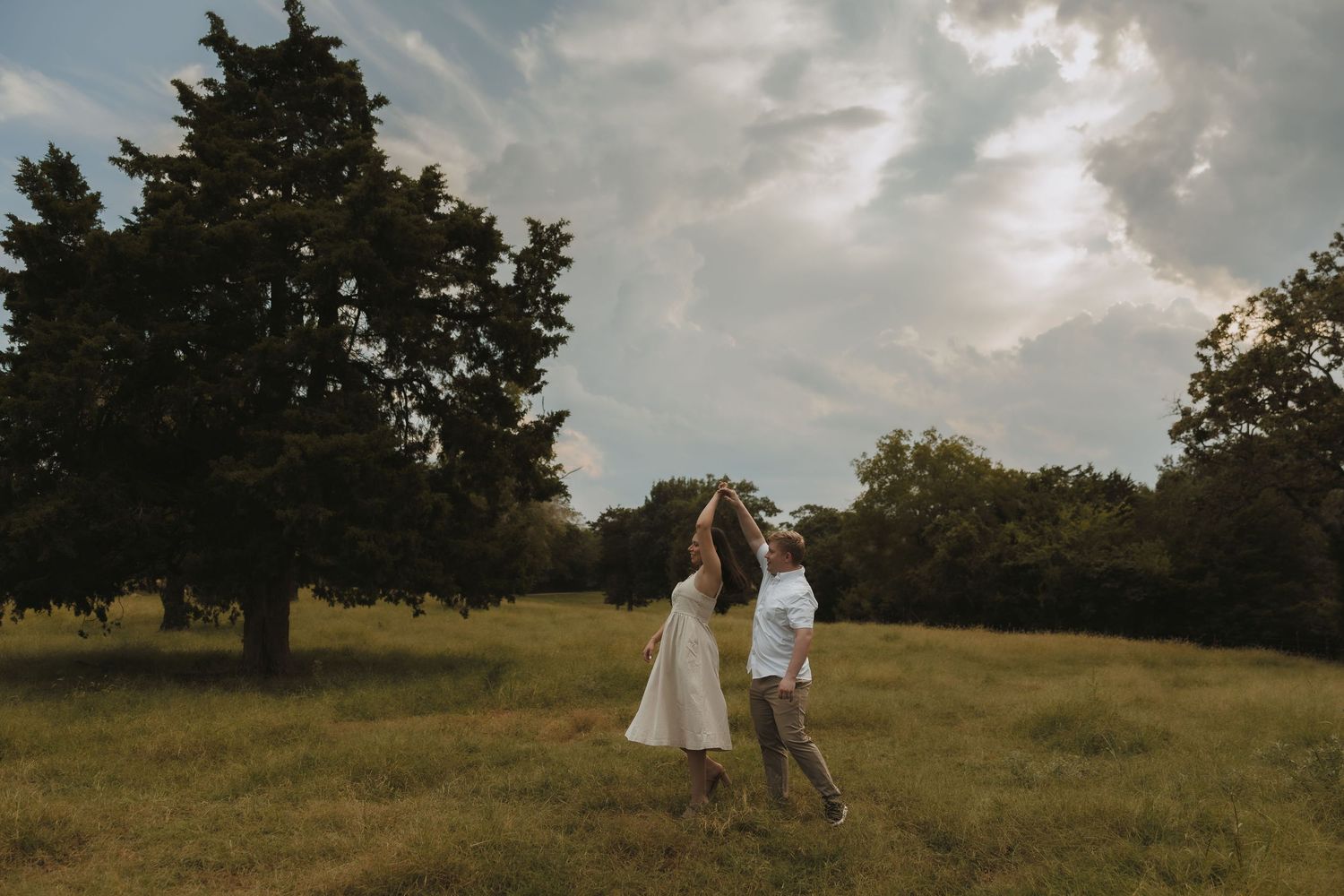 A couple dances joyfully in a lush green meadow under dramatic cloudy skies. Tulsa Photographer