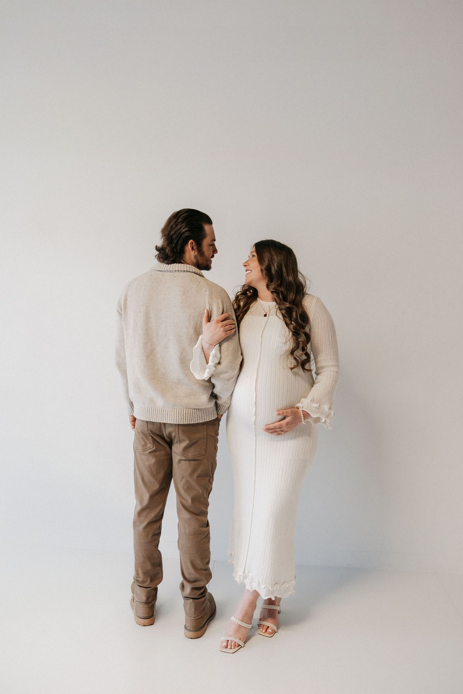Pregnant couple in neutral tones facing each other tenderly in a bright studio setting. Tulsa Photographer
