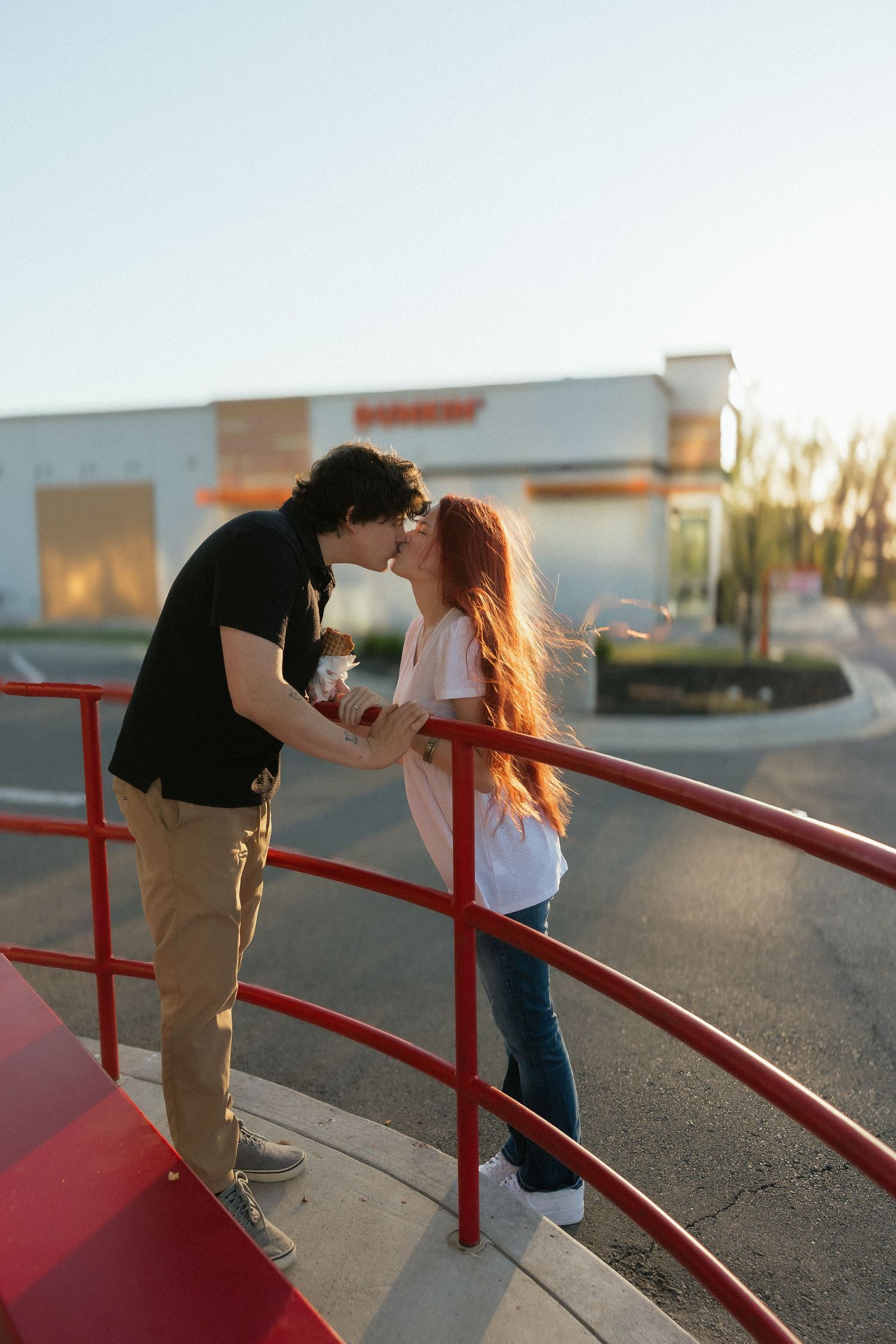 A young couple shares a romantic kiss on red railings outside a Dunkin restaurant at sunset. Tulsa Photographer