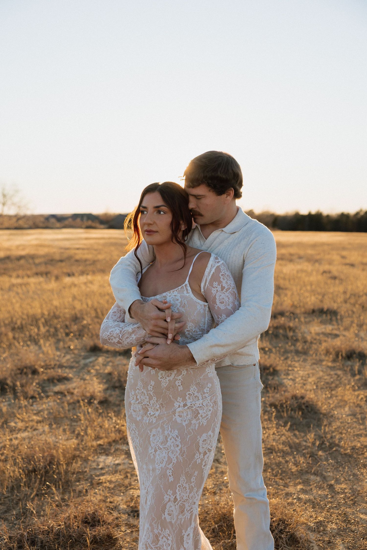 Bride in lace gown embraced by groom in white suit during golden hour field portrait. Tulsa Photographer