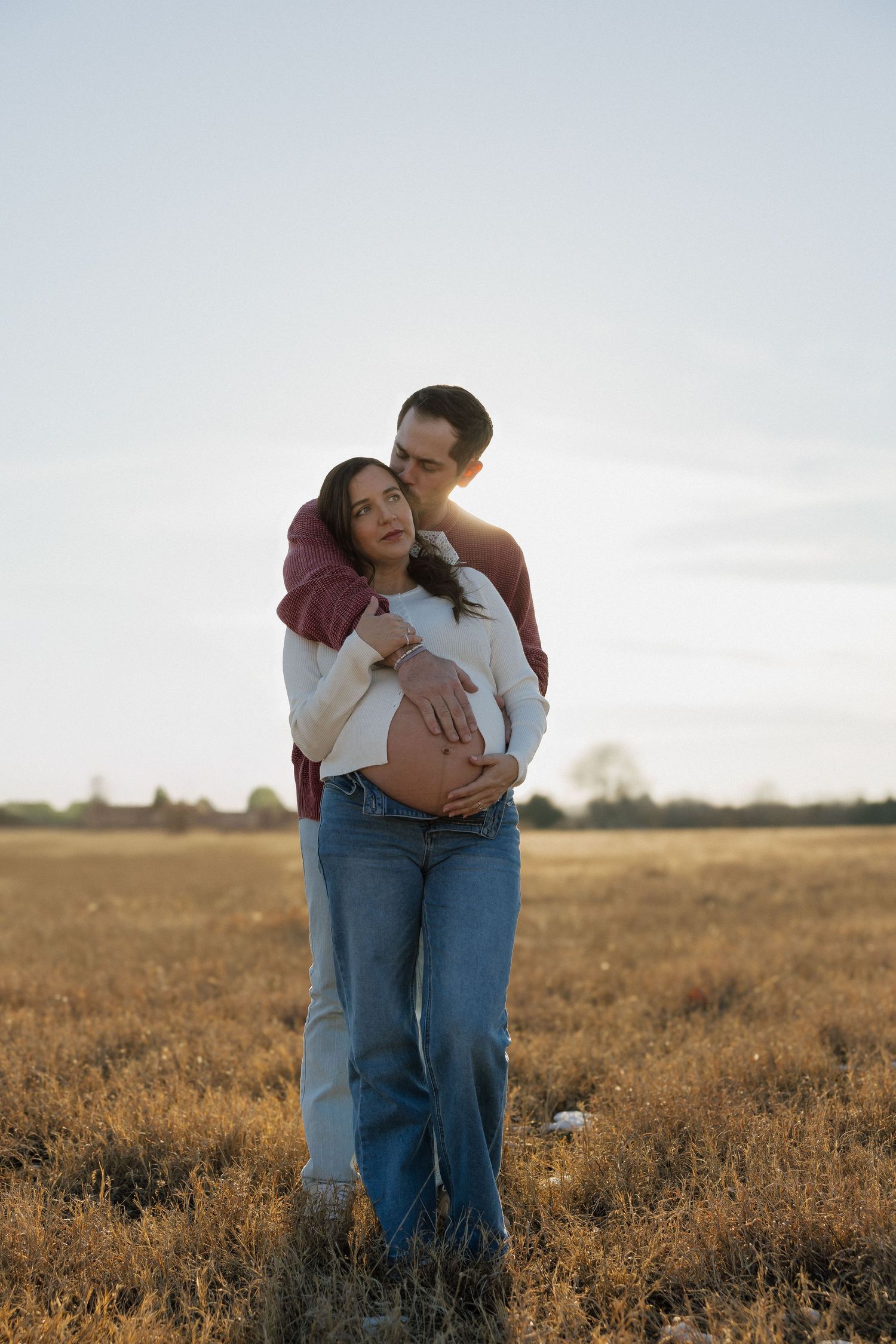 A couple embraces in a golden field at sunset, with the man holding his pregnant partner's belly tenderly.