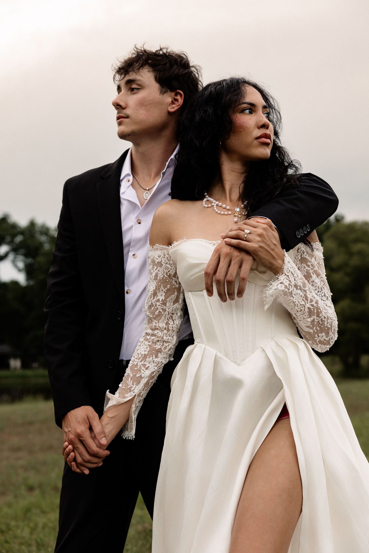 Couple in wedding attire, groom in black suit, bride in white lace corset gown with slit, posing outdoors.