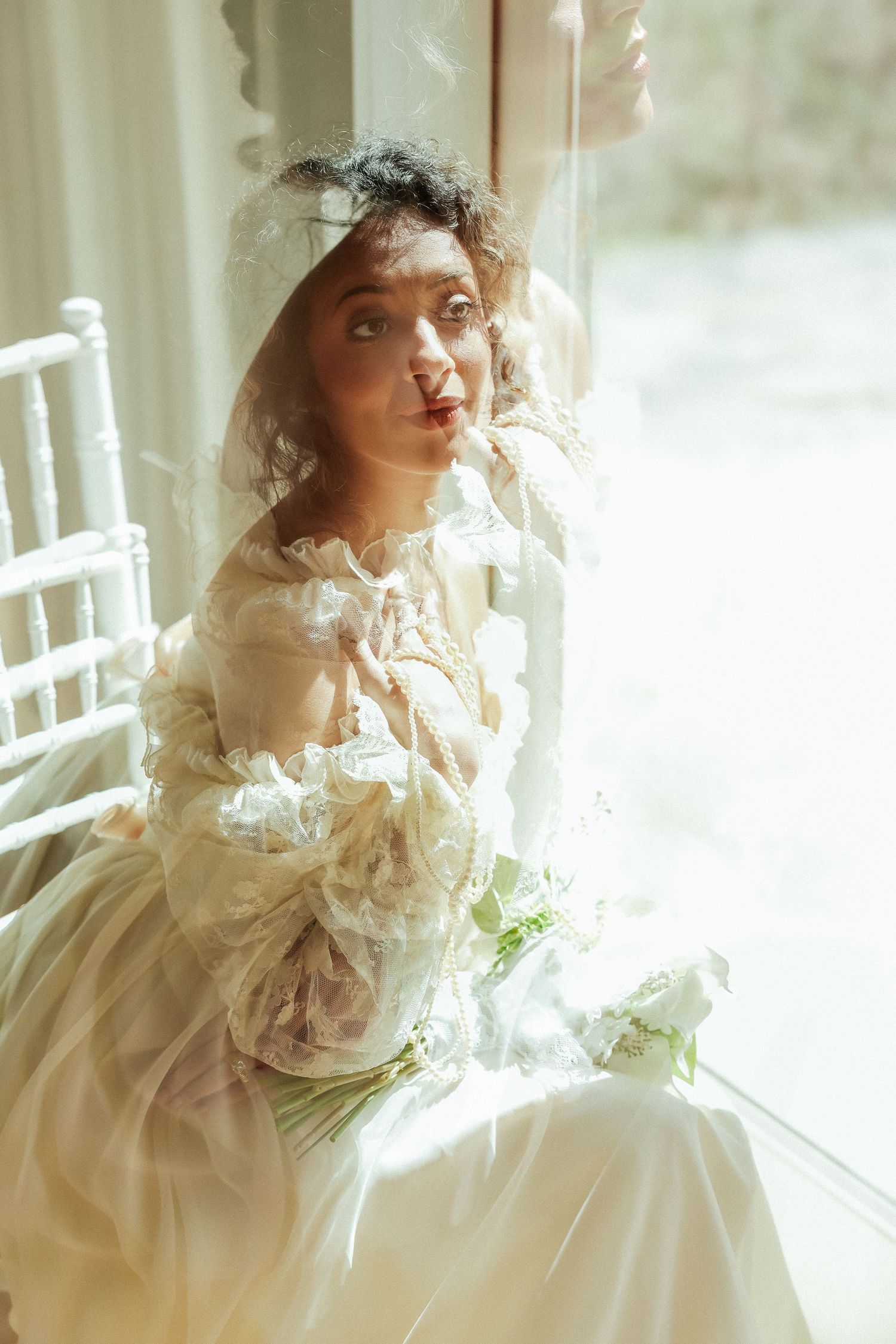 Bride in vintage lace gown with veil holds white bouquet seated by bright window in dreamy double exposure photo.