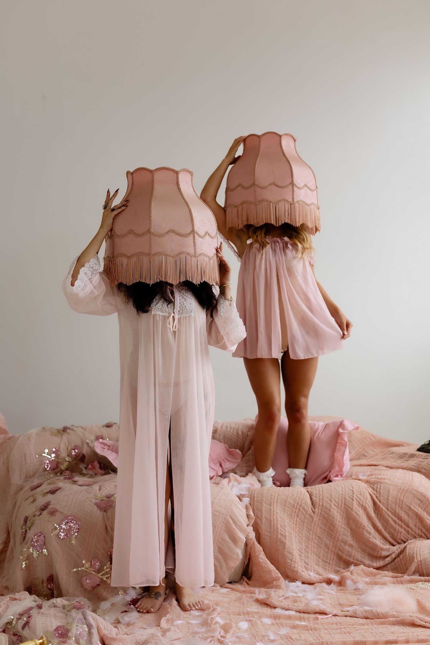 Two women standing on a bed with pink Victorian lampshades over their heads, wearing matching pink outfits. Tulsa Photographer