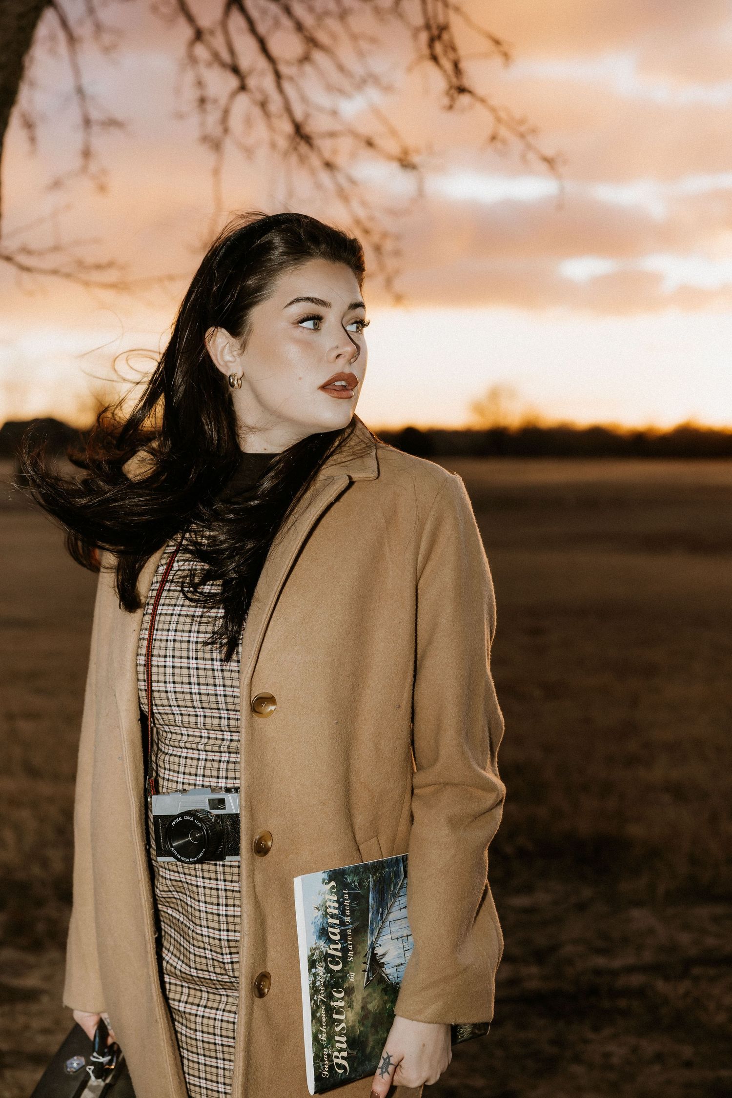 A dark-haired woman in a camel coat holds a book and camera at a dramatic golden sunset outdoors. Tulsa Photographer