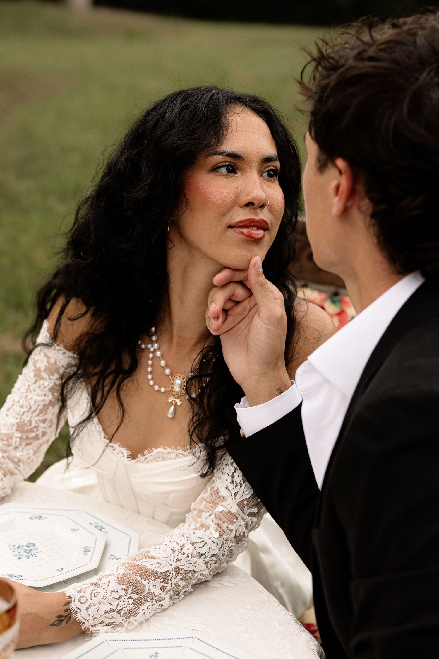 A bride in lace wedding dress and pearl necklace gazes at groom in black suit during outdoor reception.
