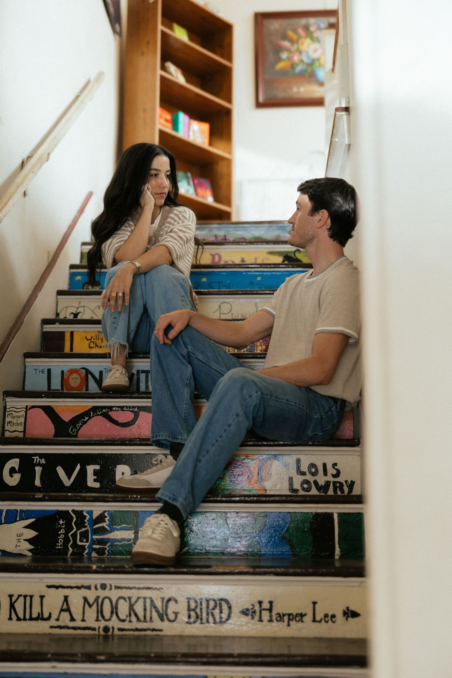 Couple sitting on book-themed painted stairs featuring Kill a Mockingbird and The Giver in a cozy bookstore setting.