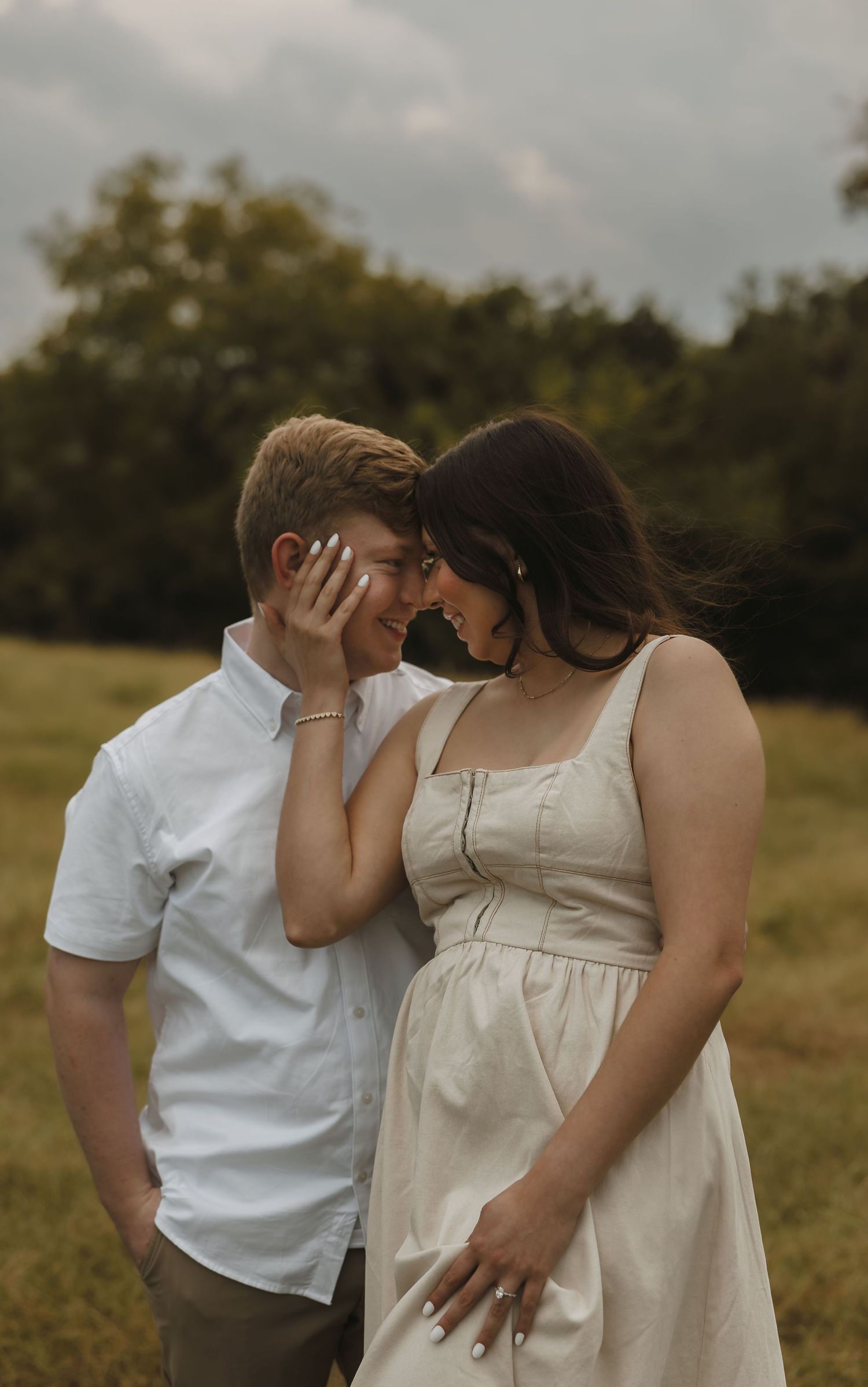 Smiling couple touching foreheads in a green field, woman in linen dress gently cupping mans face. Tulsa Photographer