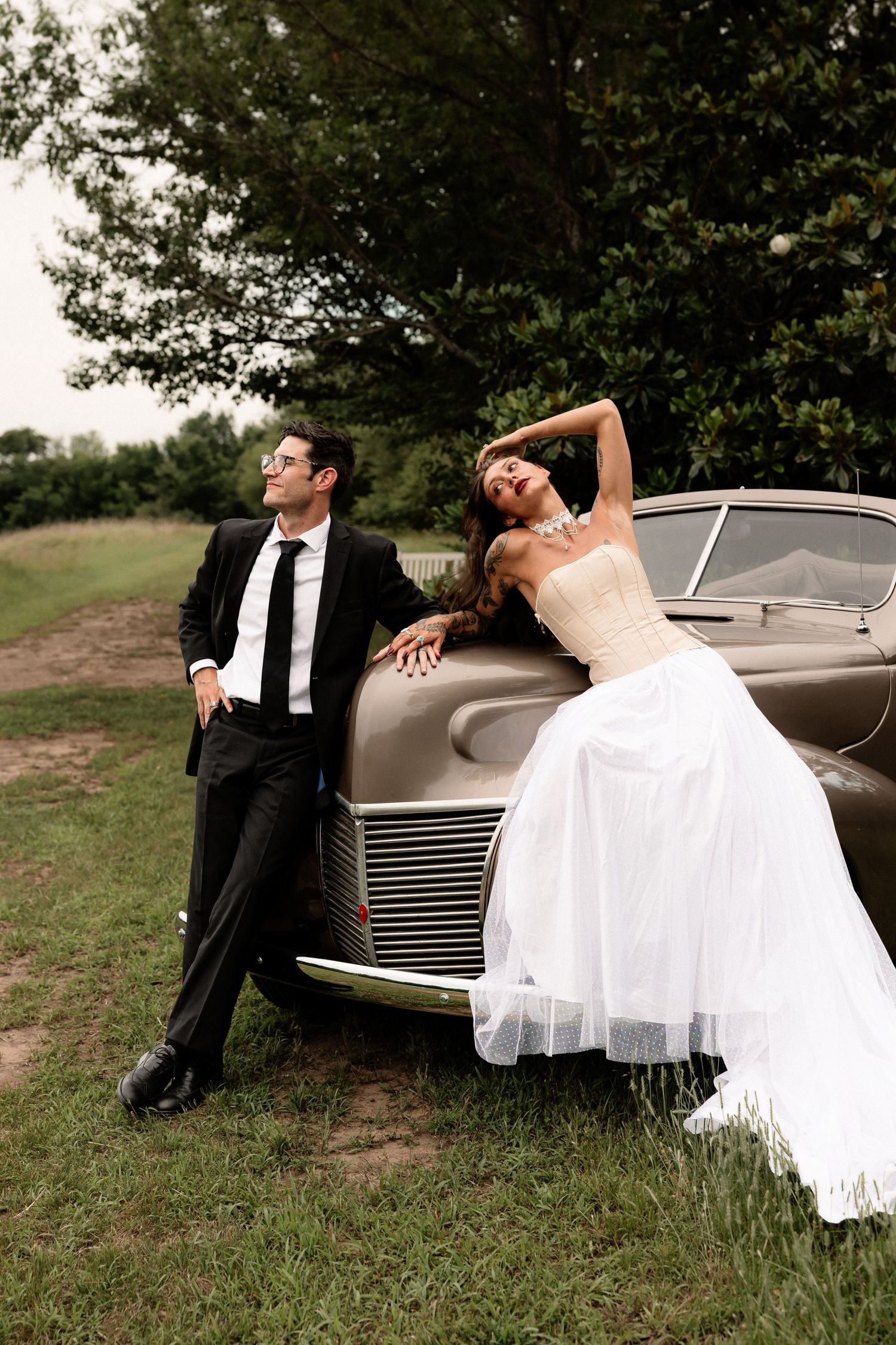 Bride in white gown and groom in black suit pose against a vintage car outdoors.