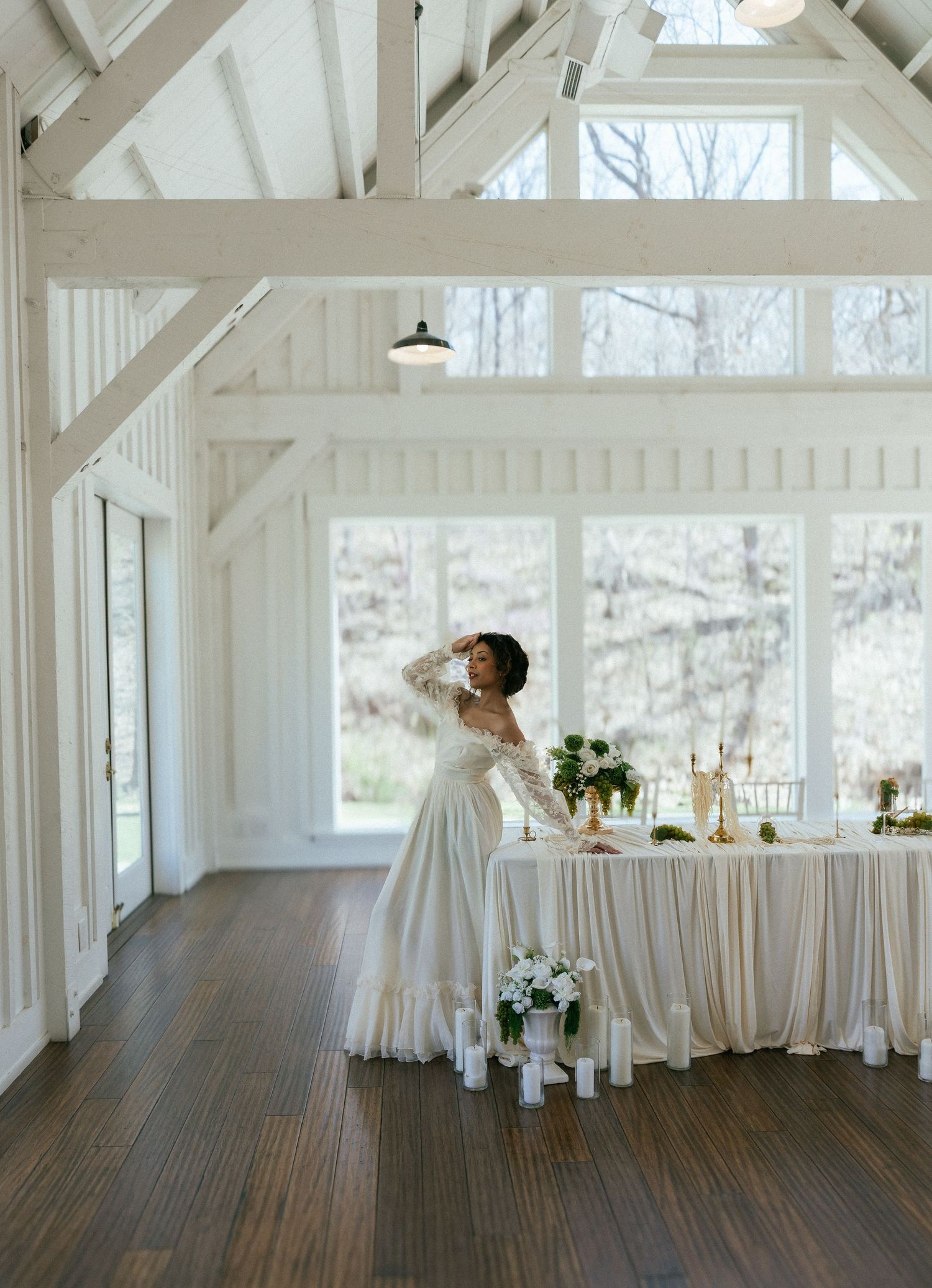 Bride in white off-shoulder gown standing in a bright white barn wedding venue with decorated table. Tulsa Photographer