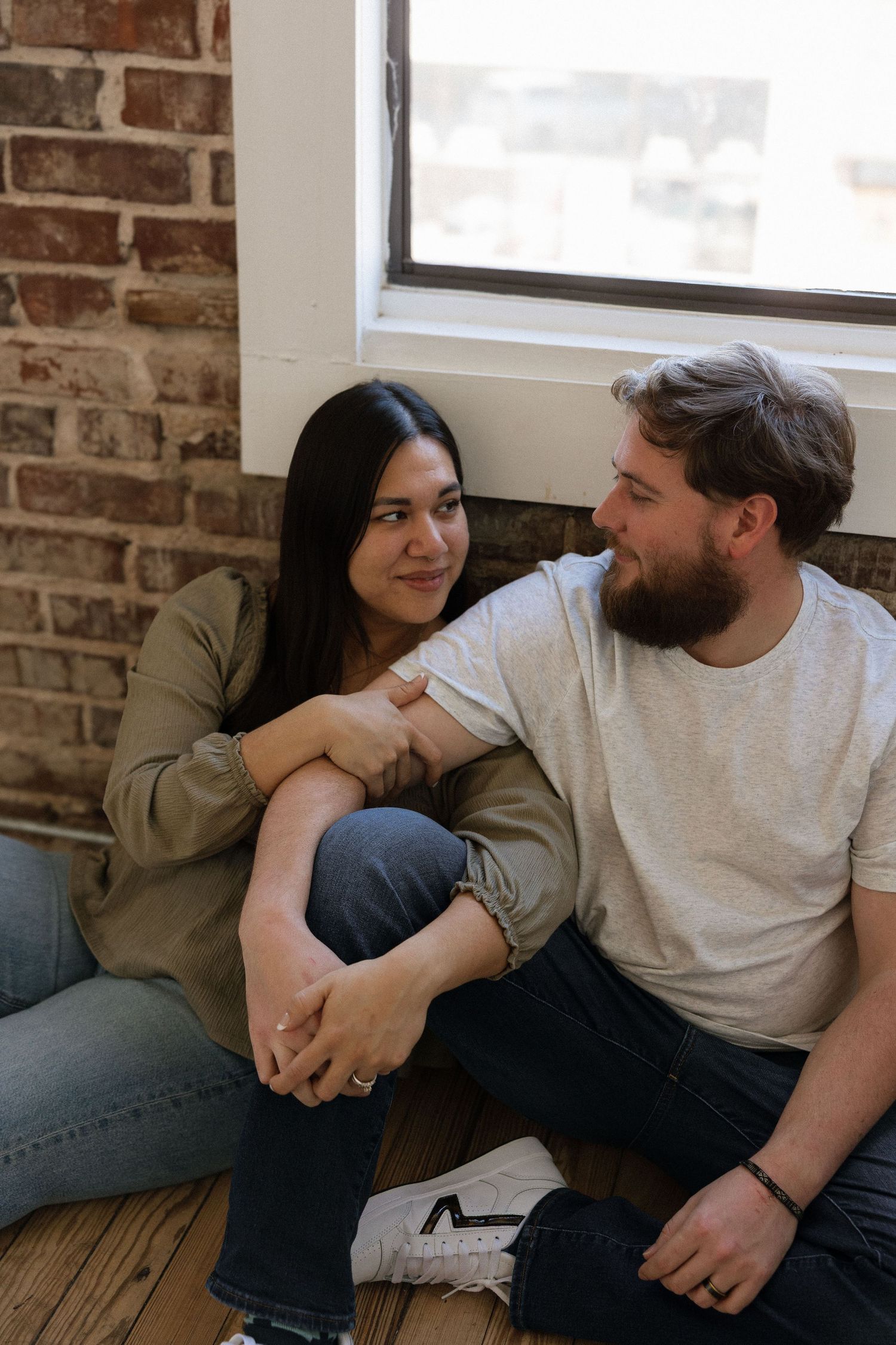 A couple sits on a wooden floor sharing a smile in a cozy loft with exposed brick and natural light. Tulsa Photographer