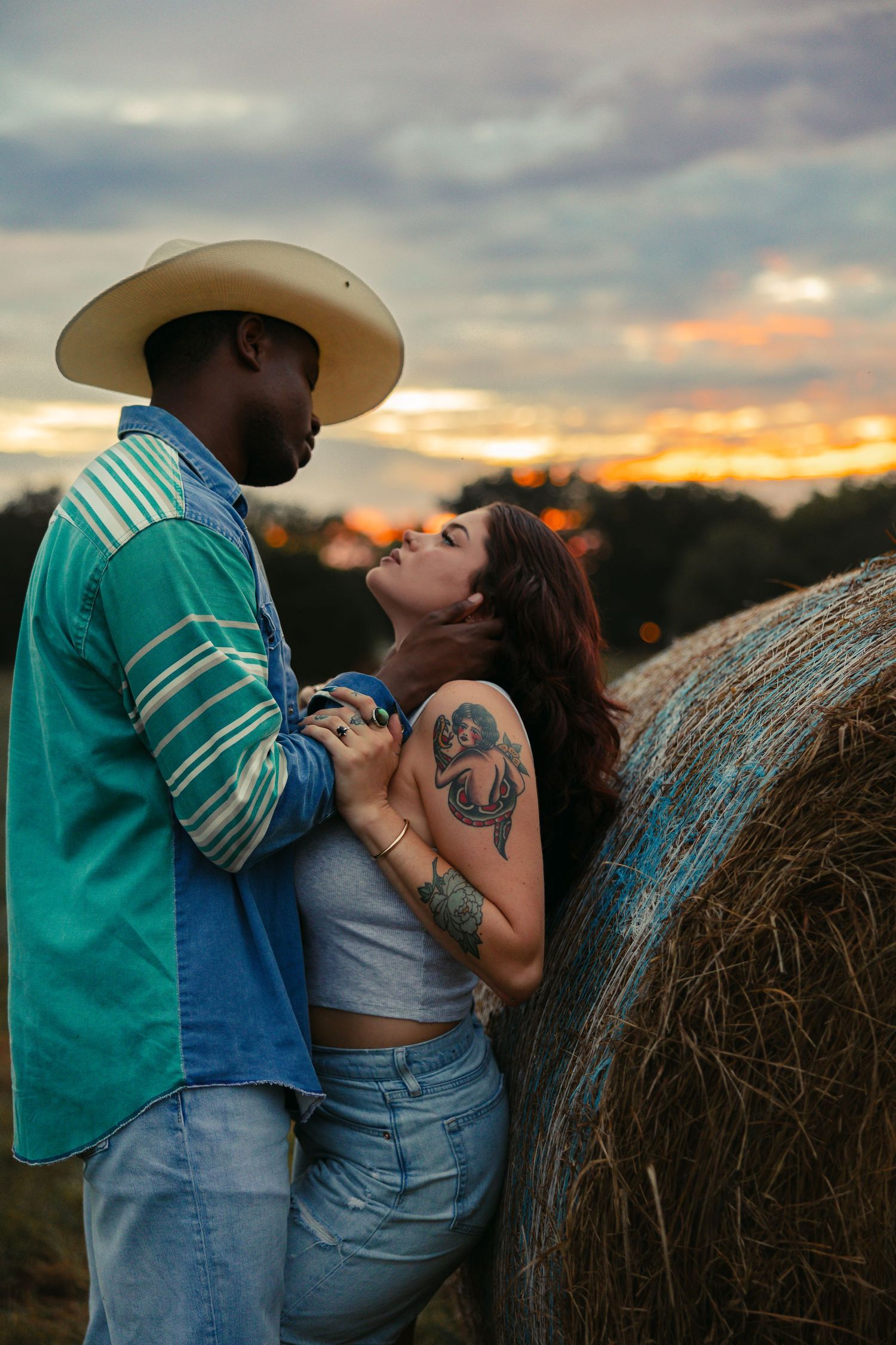Romantic interracial couple embracing near a hay bale at sunset, man in cowboy hat, woman with anchor tattoo. Tulsa Photographer