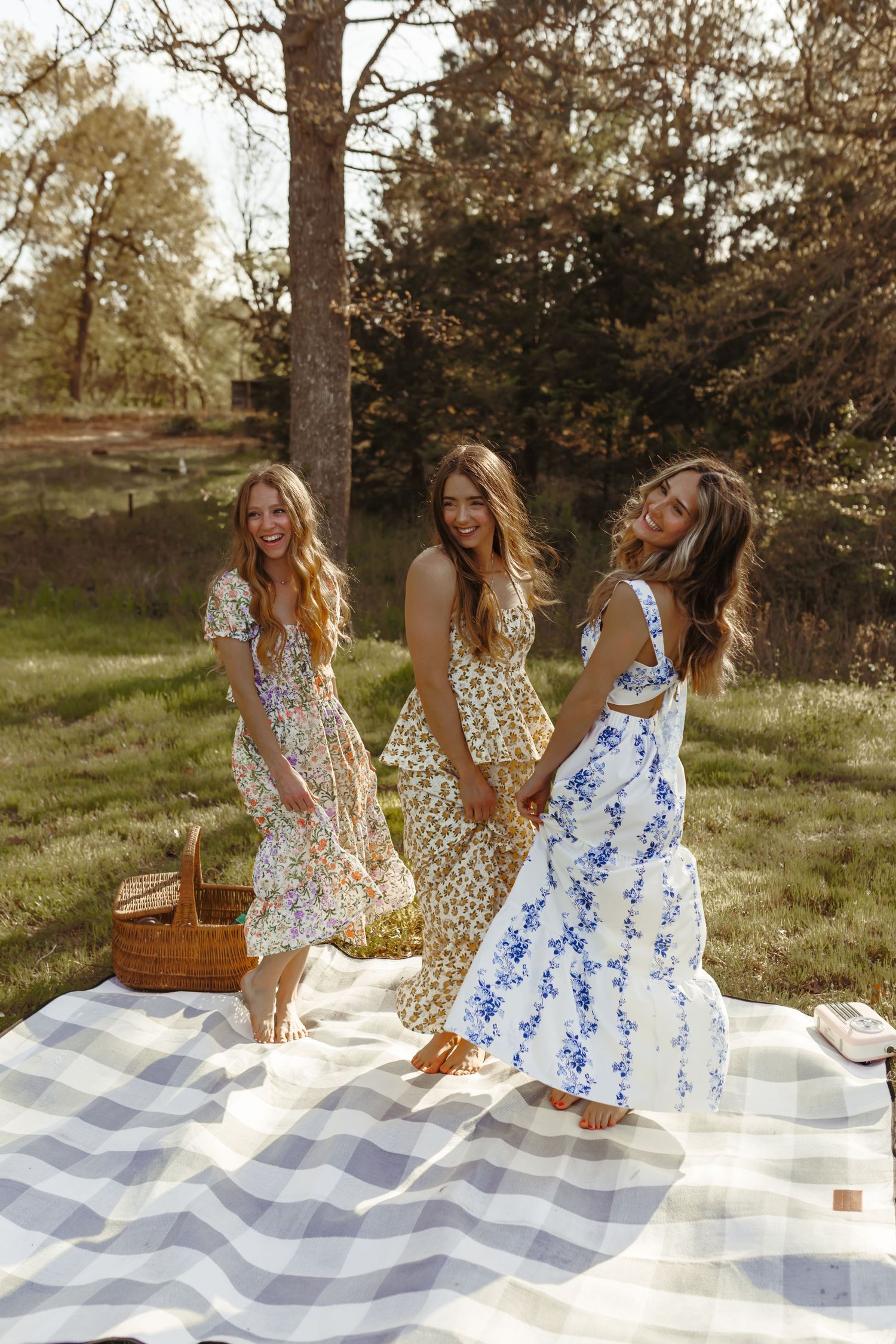 Three women in floral summer dresses dancing and holding hands on striped blanket during outdoor picnic. Tulsa Photographer