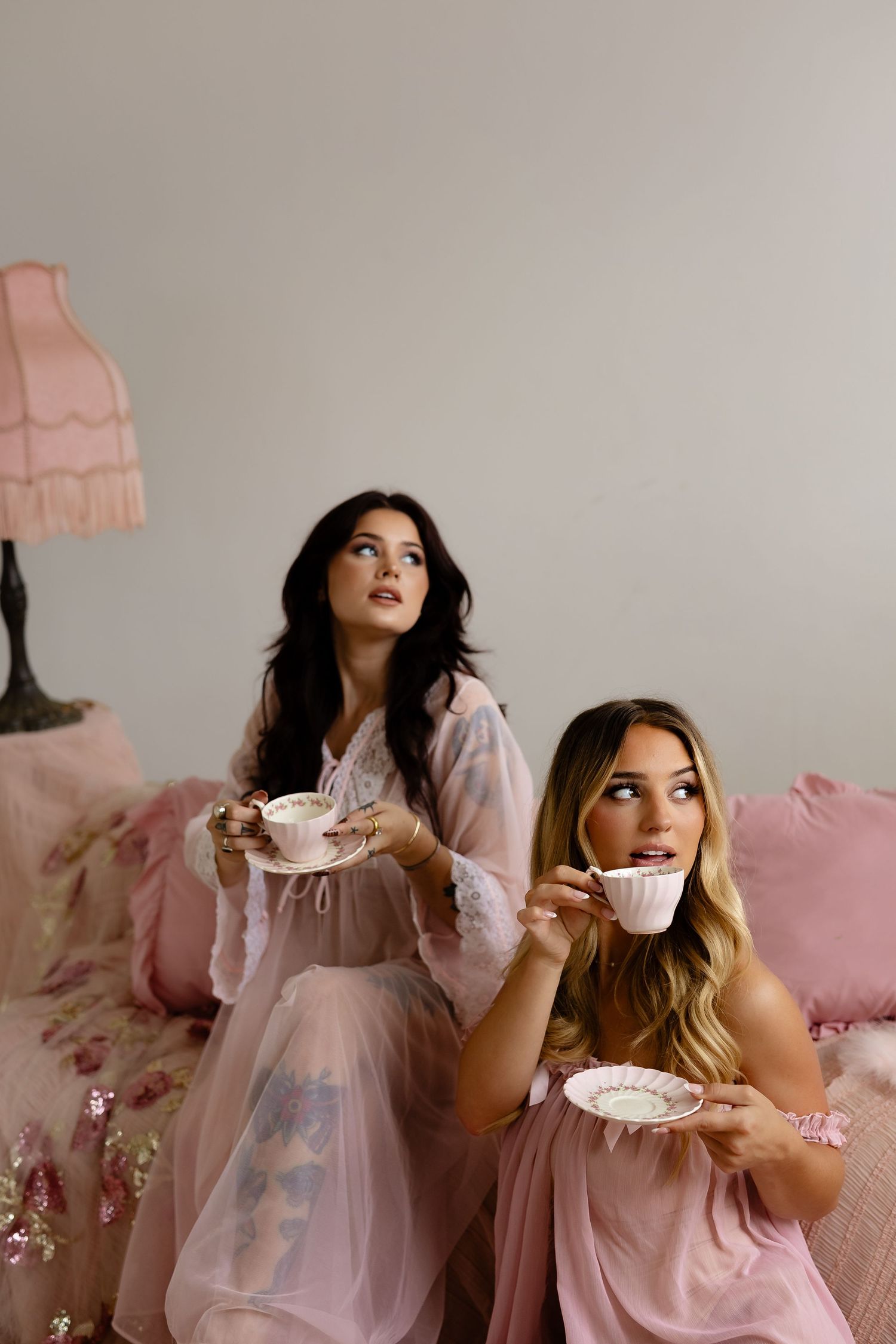 Two women in pink robes sit on a floral bed holding vintage teacups in a dreamy feminine setting. Tulsa Photographer