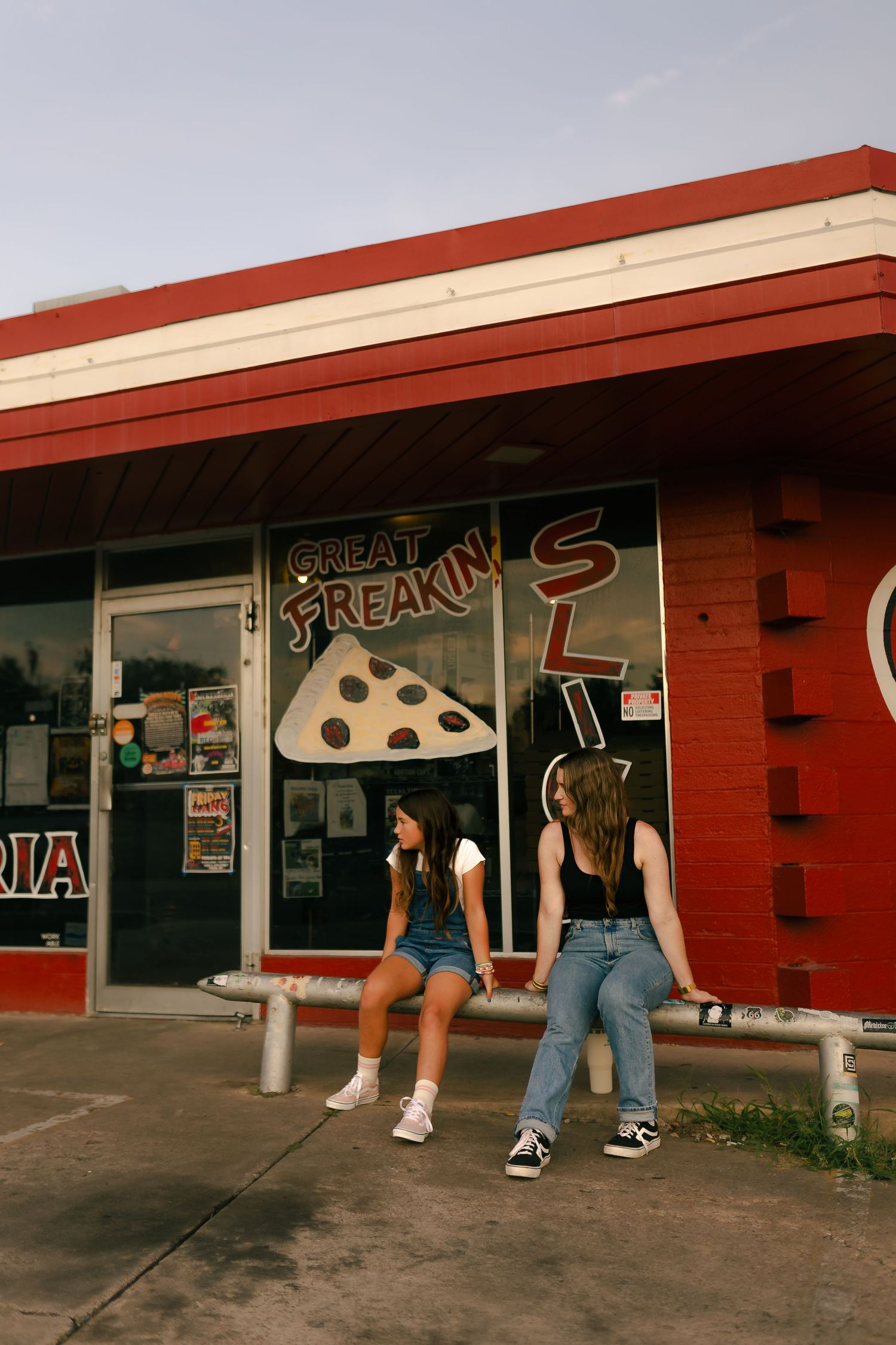 Two young women sit outside a retro pizza shop with a Great Freakin Slices sign, wearing casual denim outfits. Tulsa Photographer