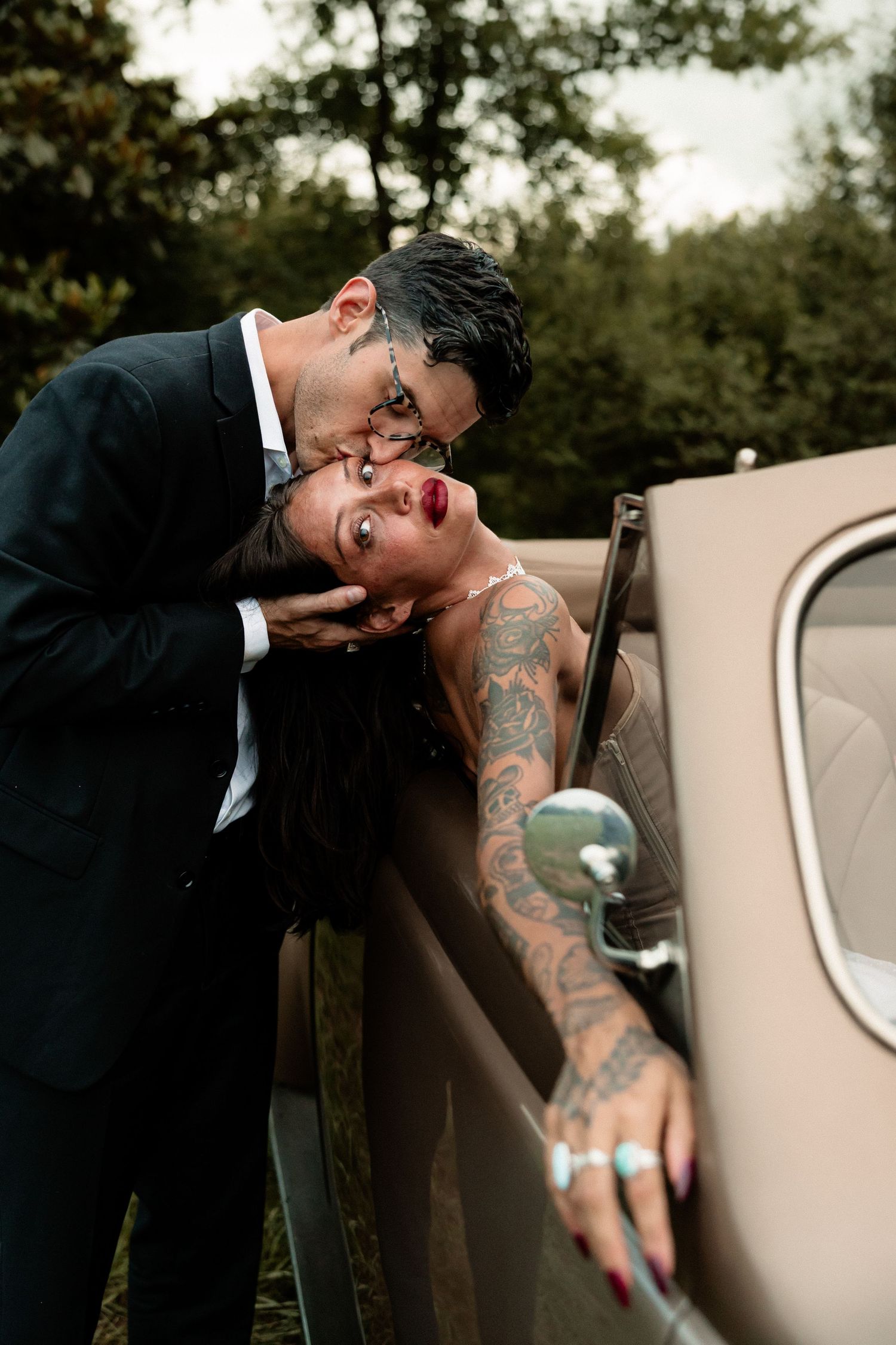 A couple sharing a romantic kiss beside a vintage beige car, the woman showing elaborate tattoos in elegant wedding attire.