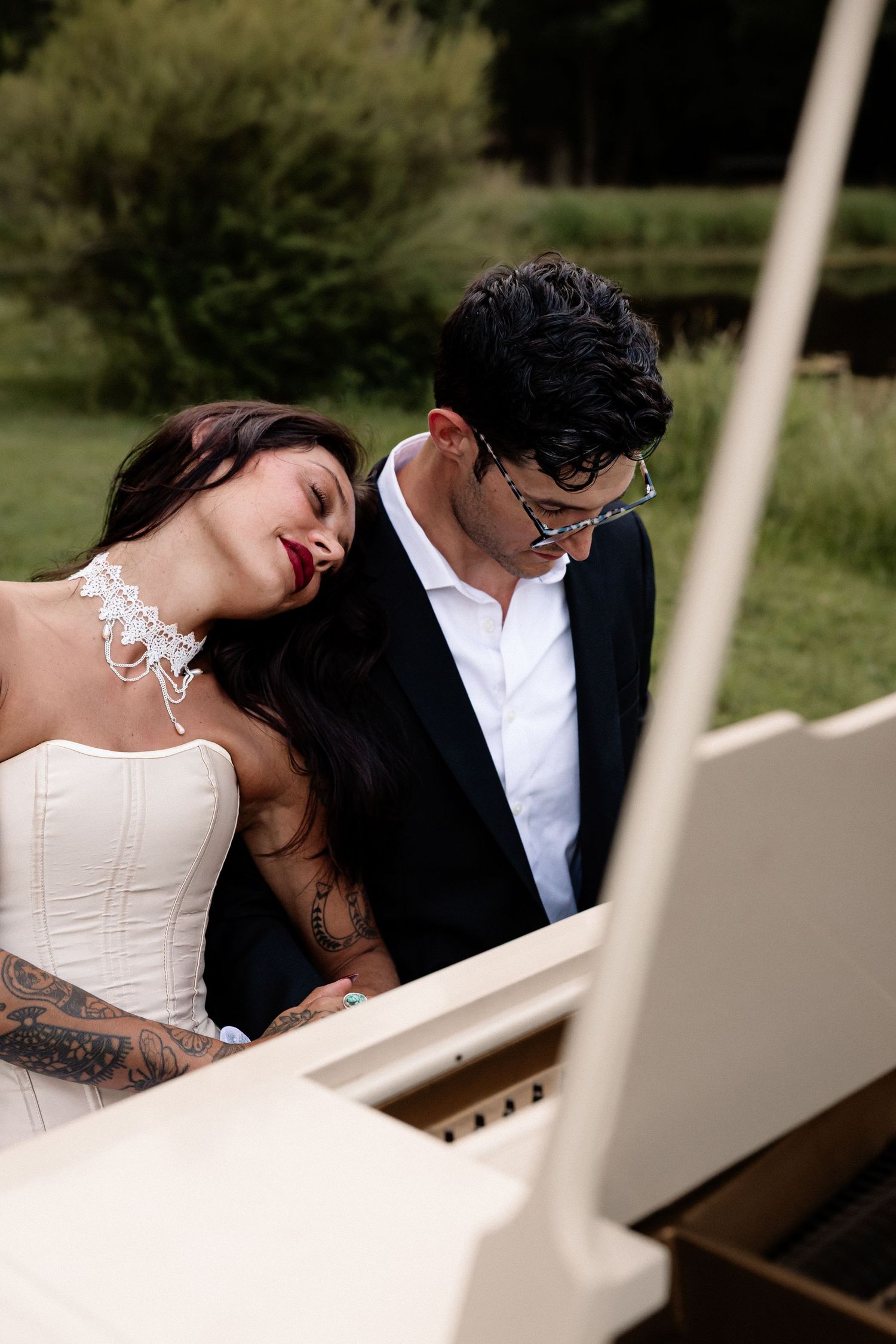 A bride and groom share a romantic moment at a white grand piano outdoors, surrounded by lush greenery.