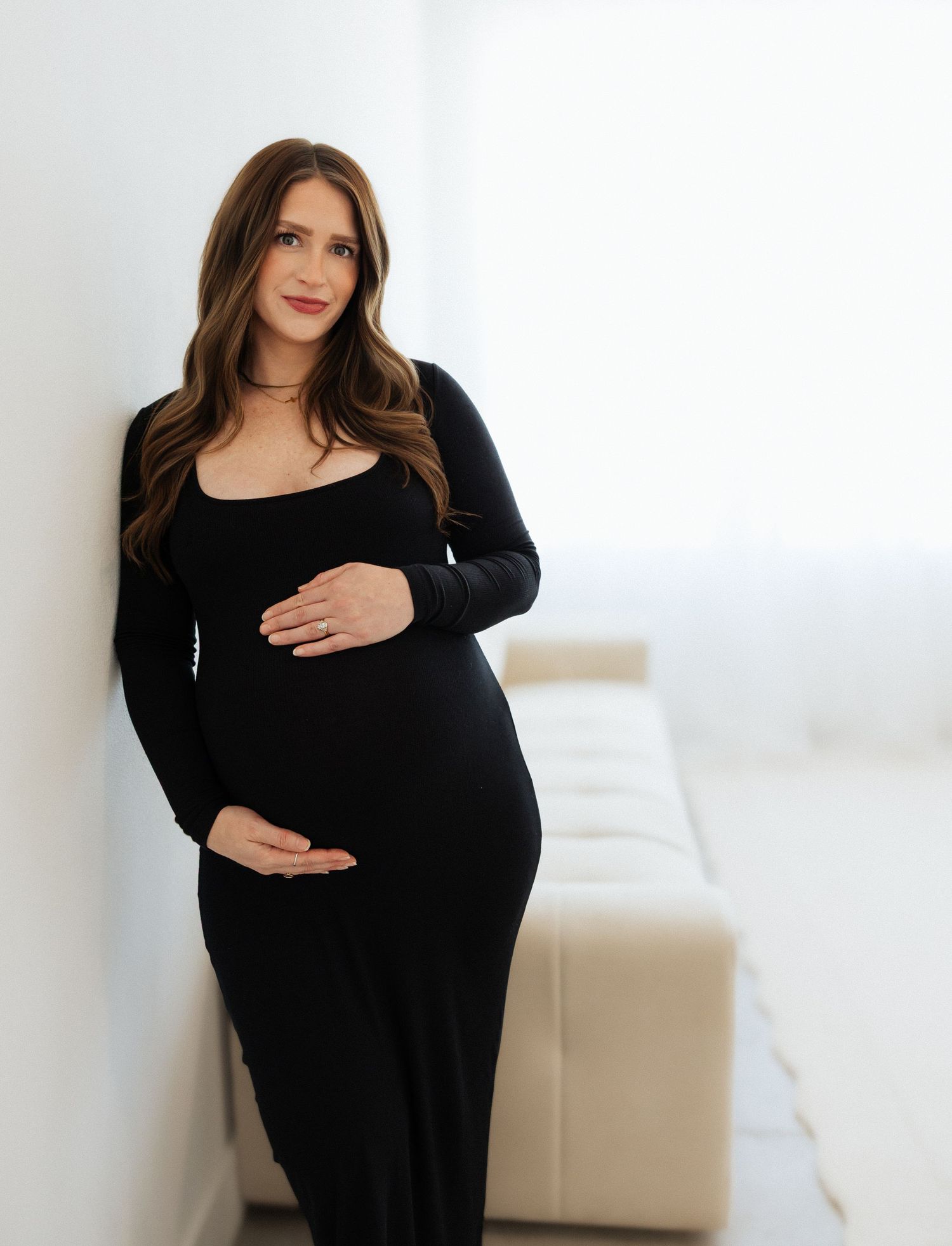 Pregnant woman in black long-sleeve dress cradling baby bump, leaning against white wall in bright studio.