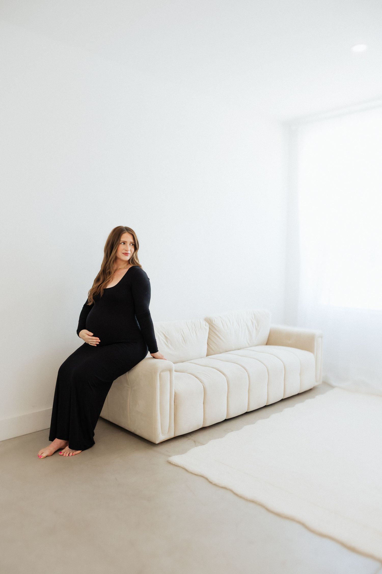 Pregnant woman in black dress sitting on cream sofa in minimalist white room.