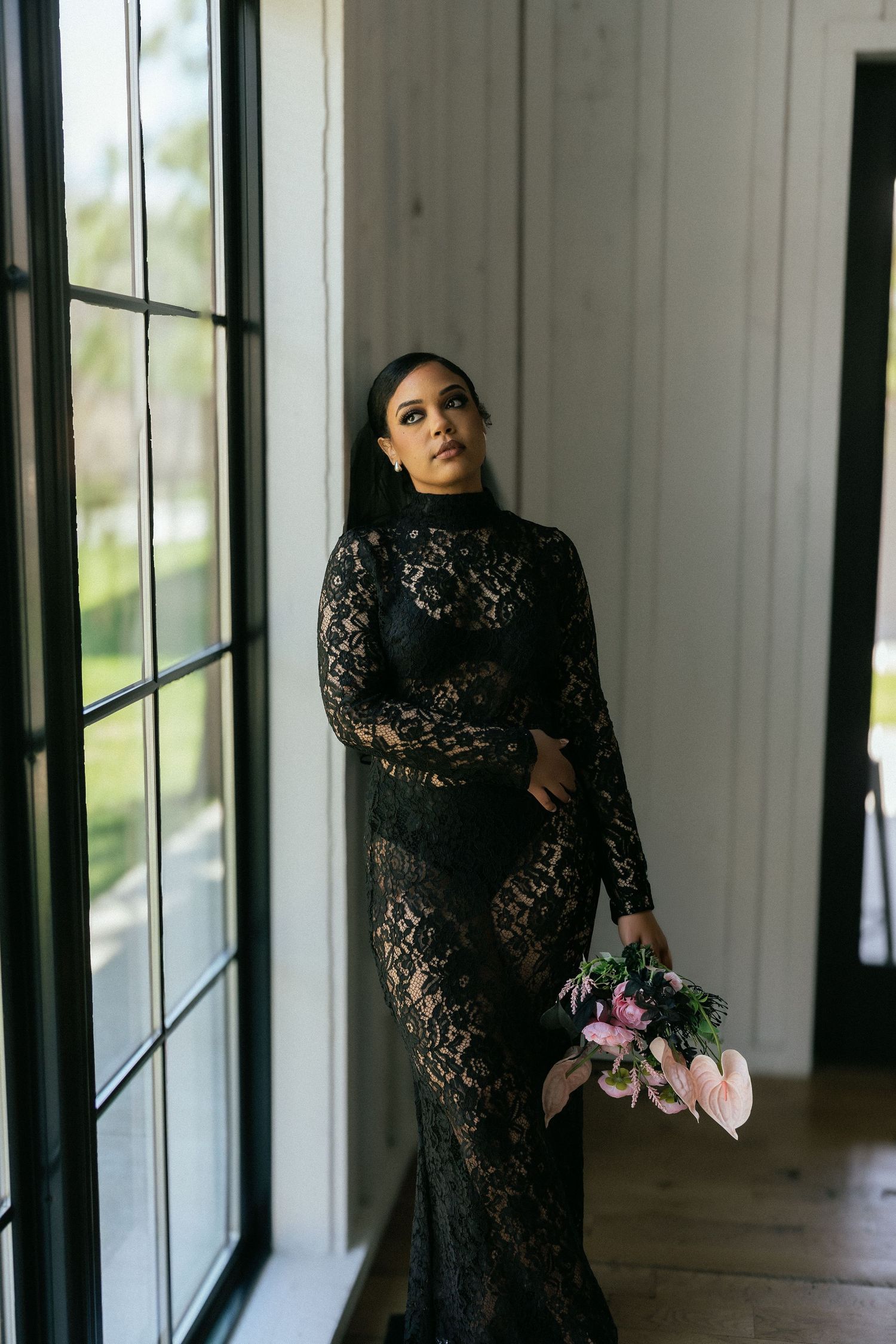 Elegant bride in black lace long-sleeve gown holding pink bouquet beside tall white windows. Tulsa Photographer