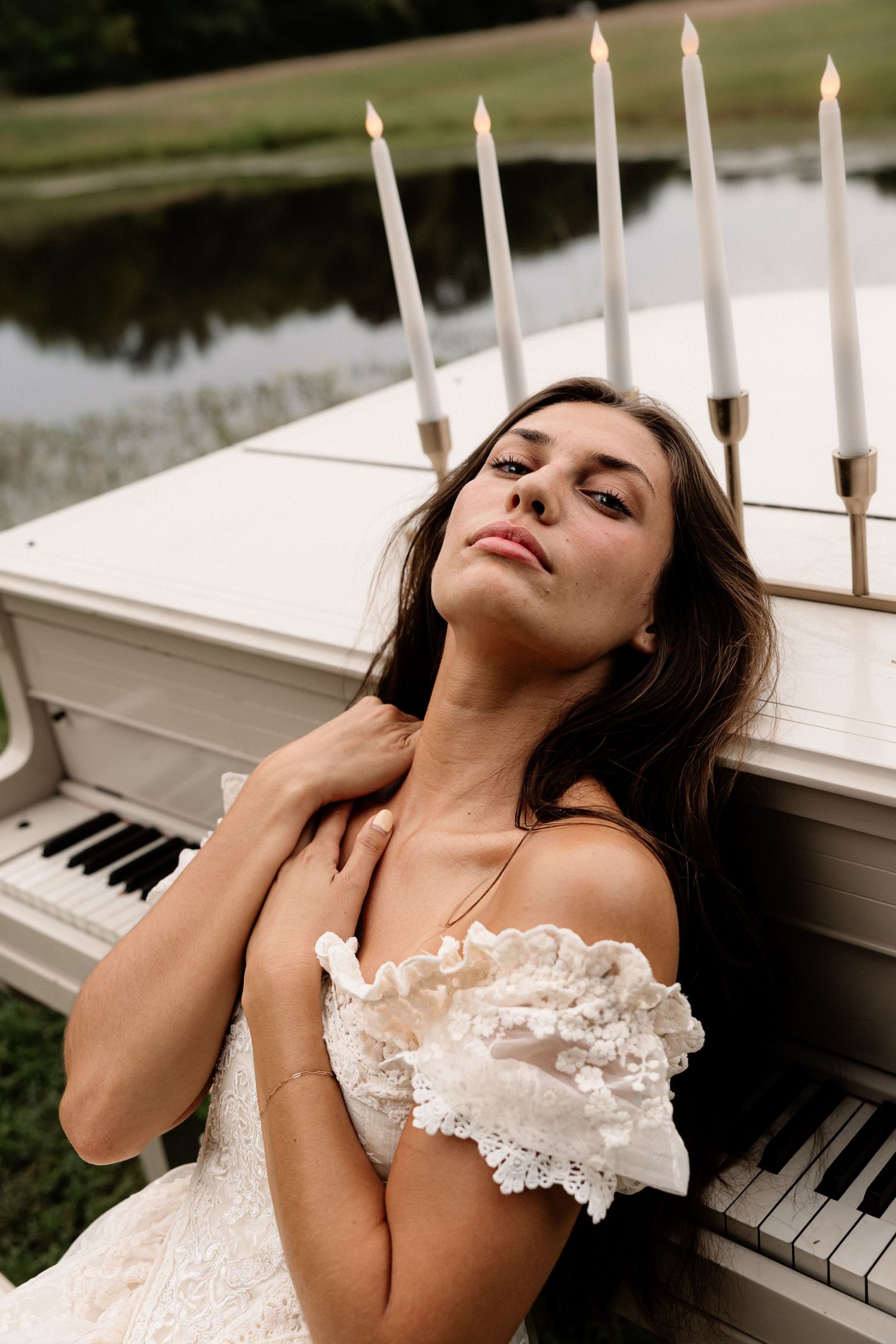 Elegant bride in white lace off-shoulder gown leaning against a white piano with candles outdoors.
