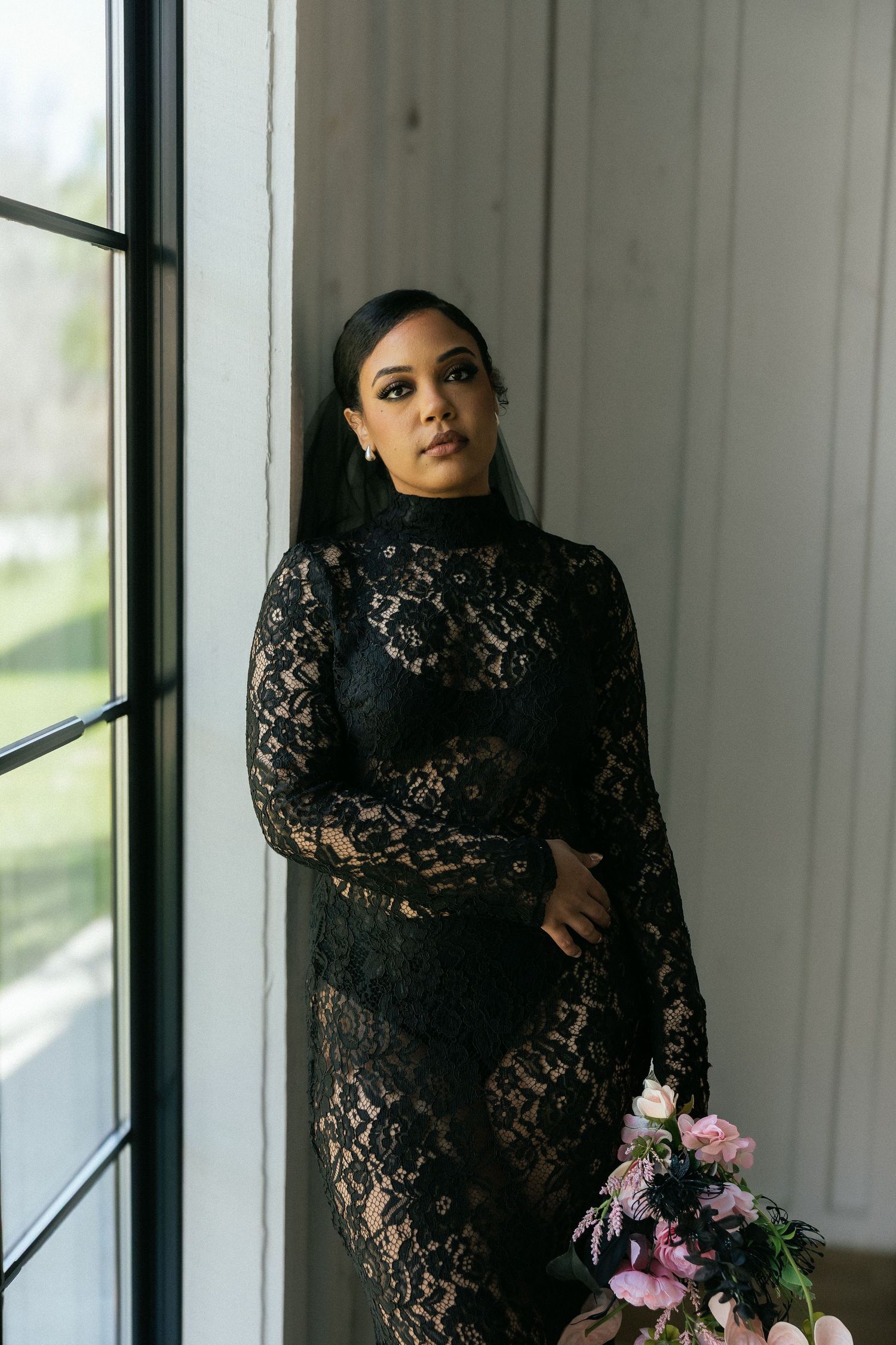 Elegant woman in black lace long-sleeve gown holding pink bouquet while standing by large window indoors. Tulsa Photographer