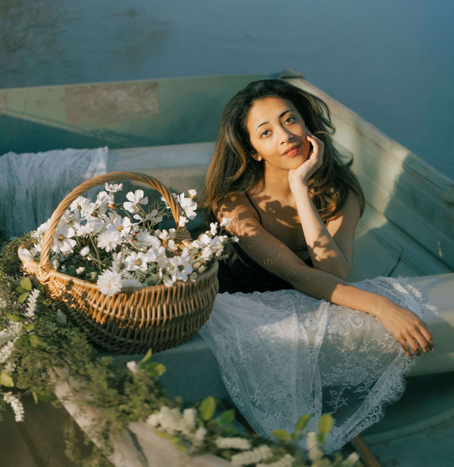 Young woman in white lace skirt reclines in a boat with a wicker basket of white flowers in soft natural light. Tulsa Photographer