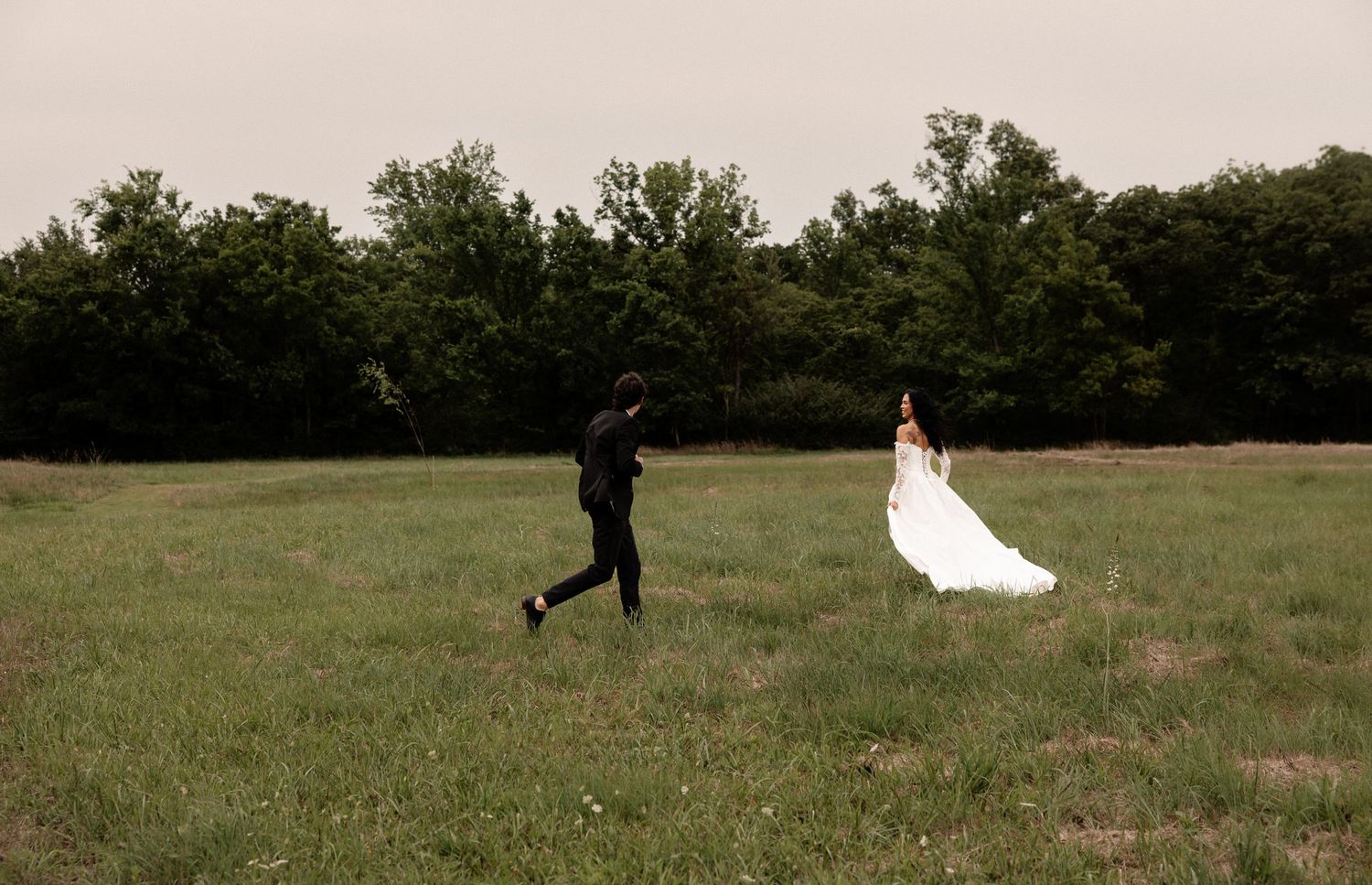 Bride in white gown and groom in black suit running through a green field on their wedding day.