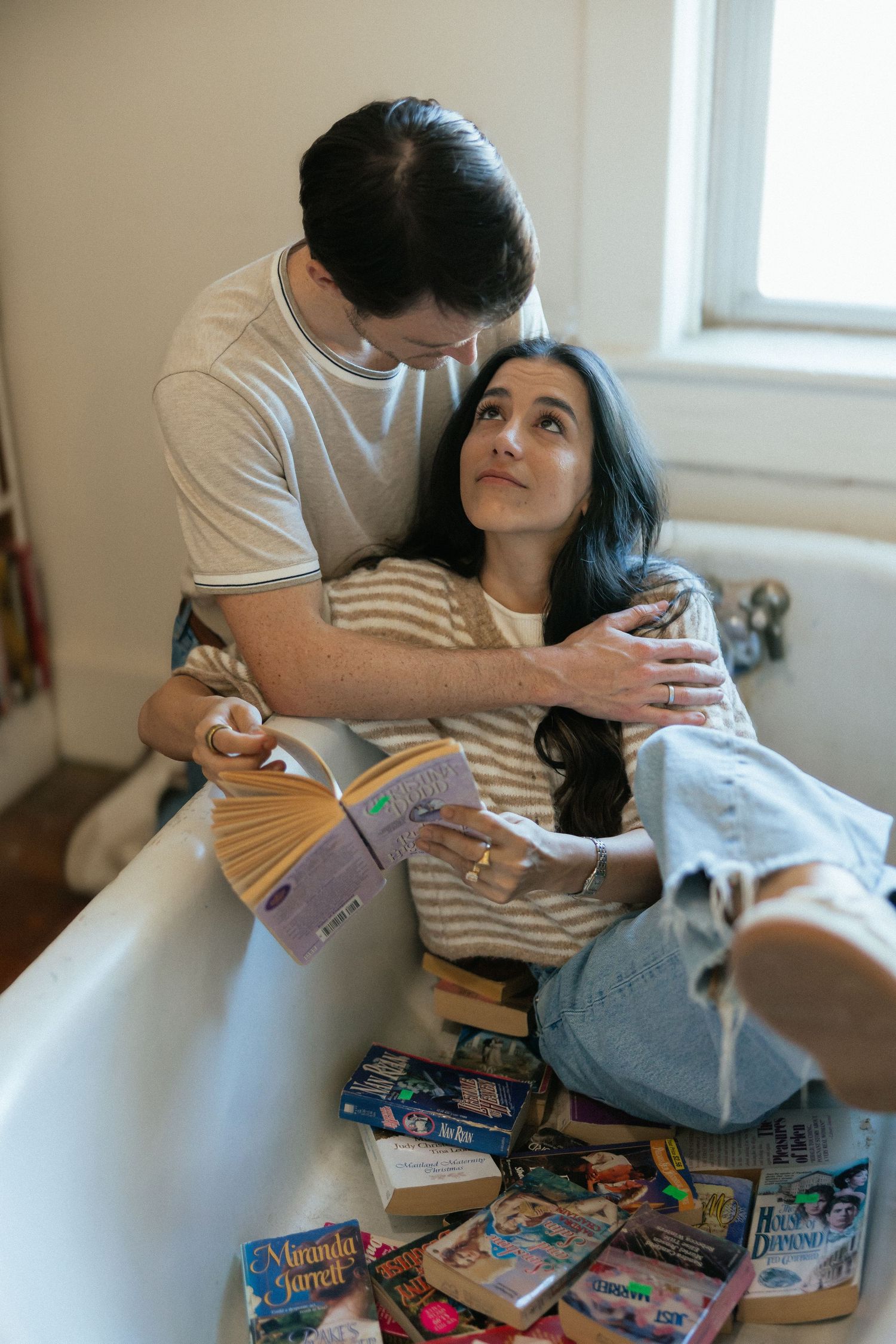 Couple sharing a tender moment in a bathtub surrounded by colorful books, woman looking up lovingly at man holding open book.