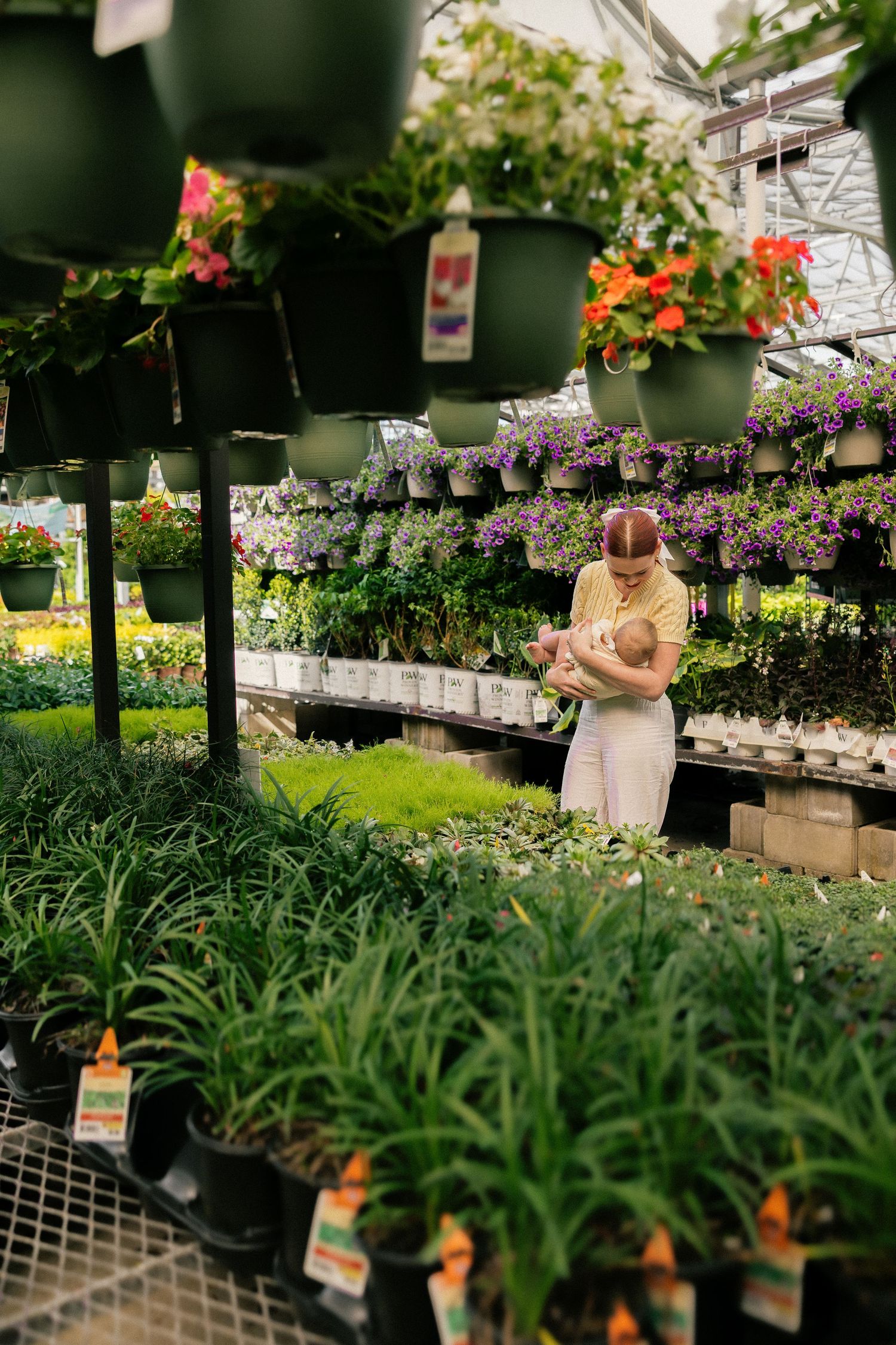 Woman holding a baby while shopping for plants in a greenhouse garden center with hanging flower baskets.
