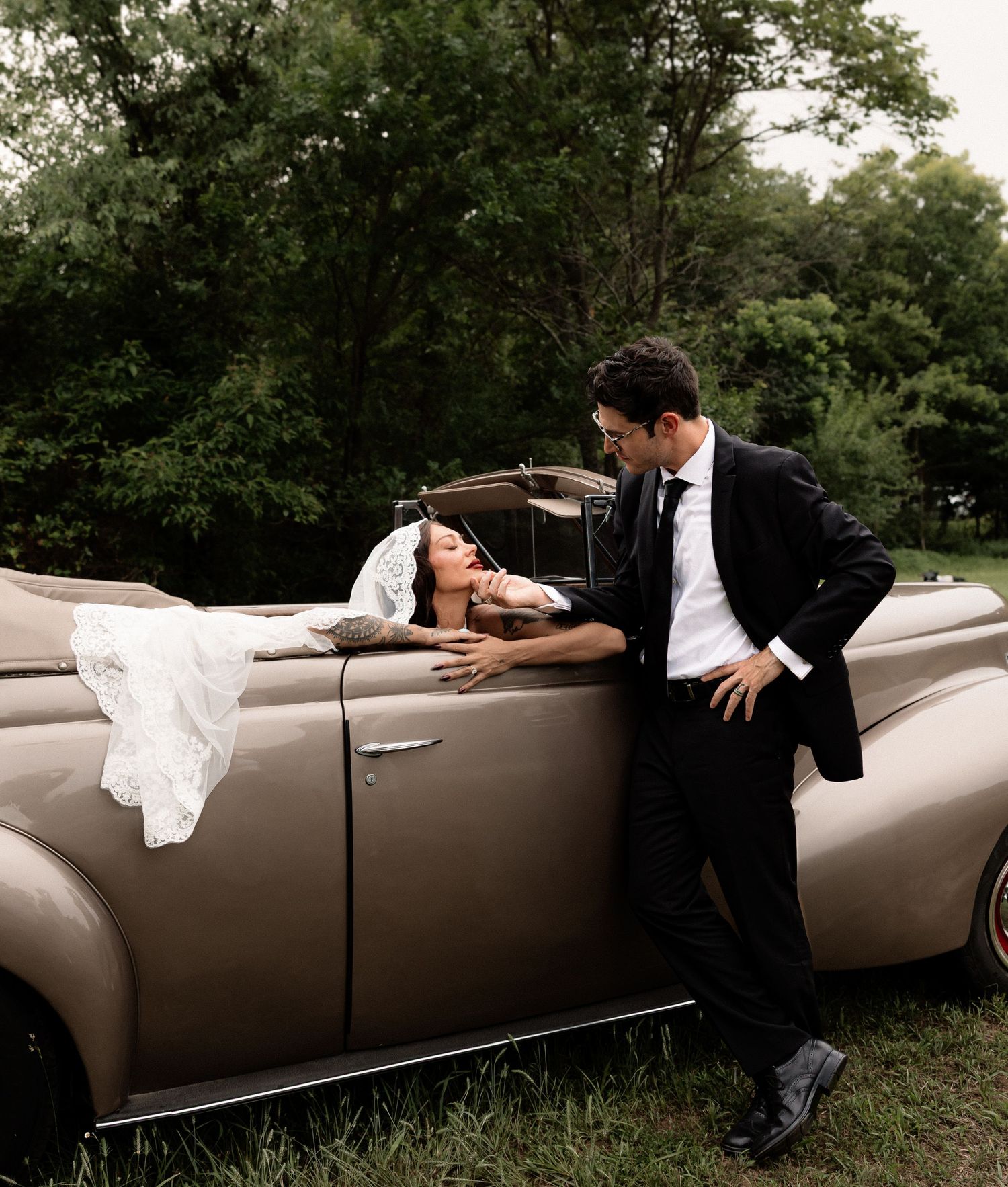 Bride and groom posing elegantly beside a vintage beige convertible car surrounded by lush green trees.