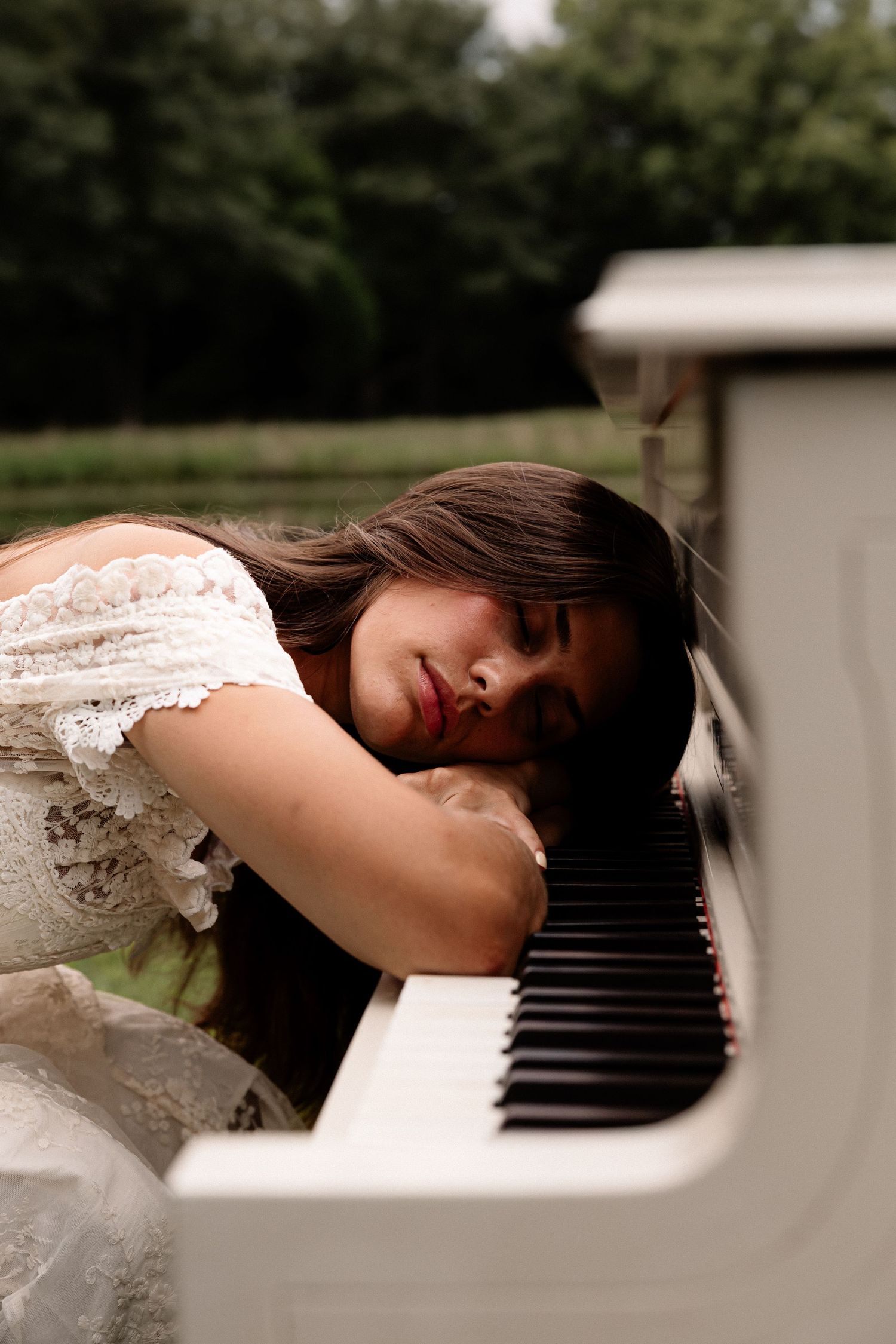 Young woman in white lace dress resting her head on white piano keys outdoors with green trees in background.