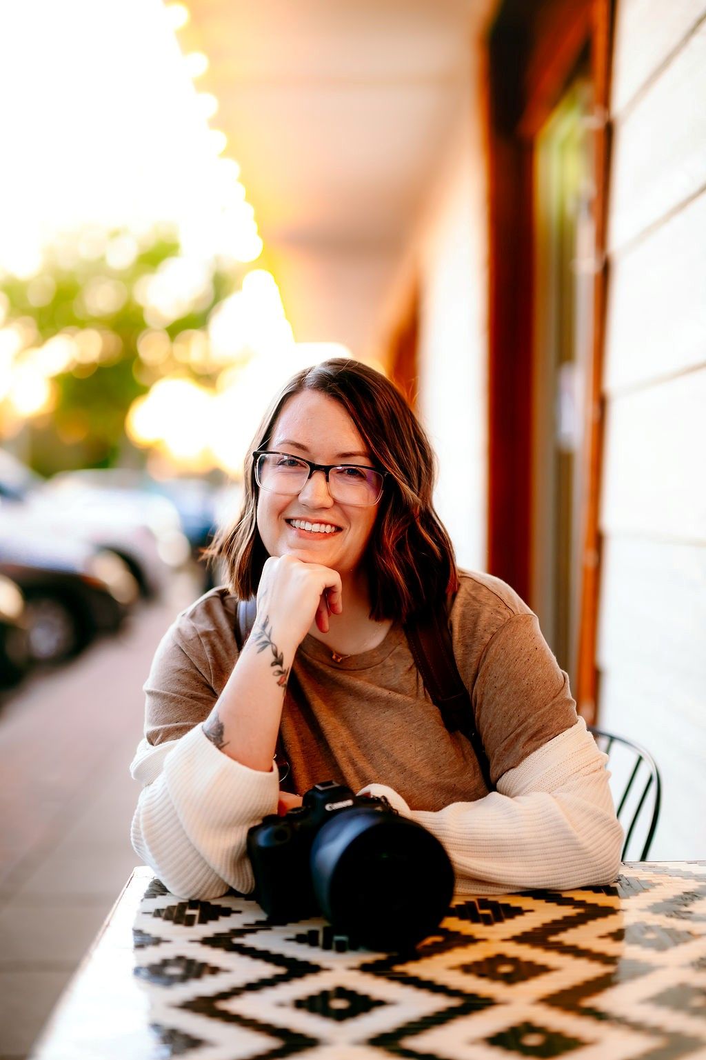Smiling female photographer with glasses sits at outdoor cafe table with camera at golden hour. Tulsa Photographer