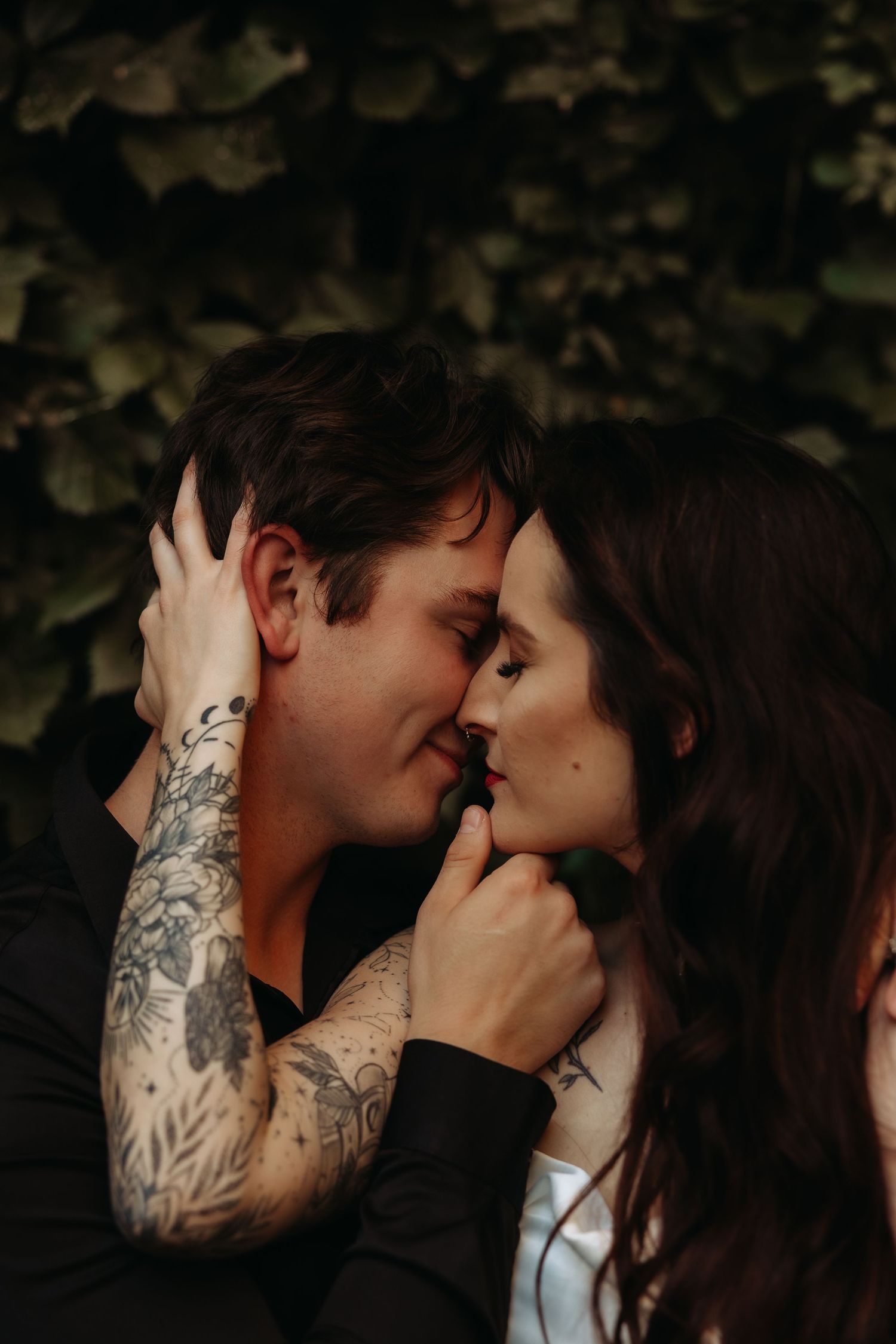 A tattooed couple shares a romantic forehead-to-forehead moment surrounded by lush green foliage.