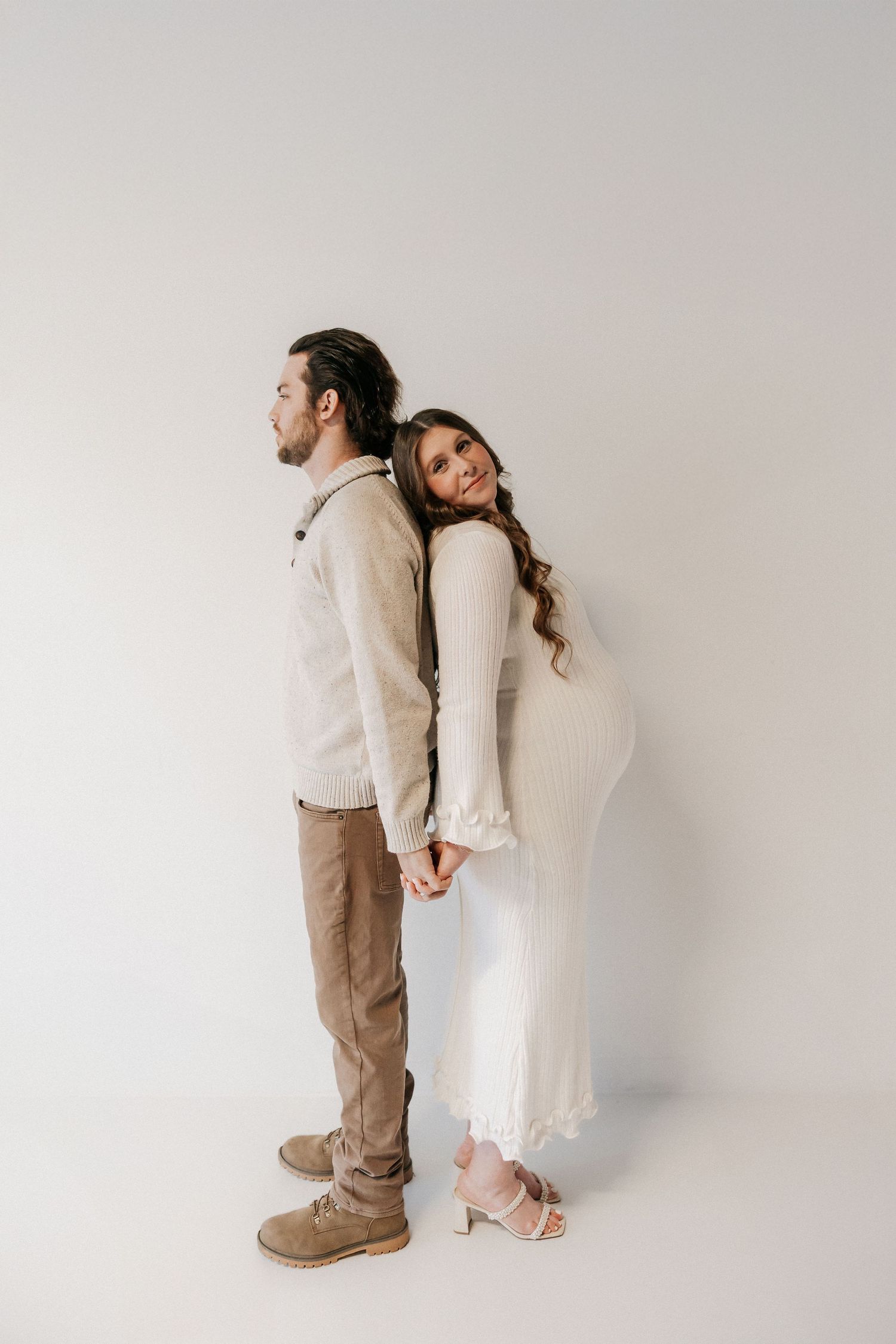 Pregnant woman in white dress holds hands with partner as they stand back-to-back in a studio. Tulsa Photographer