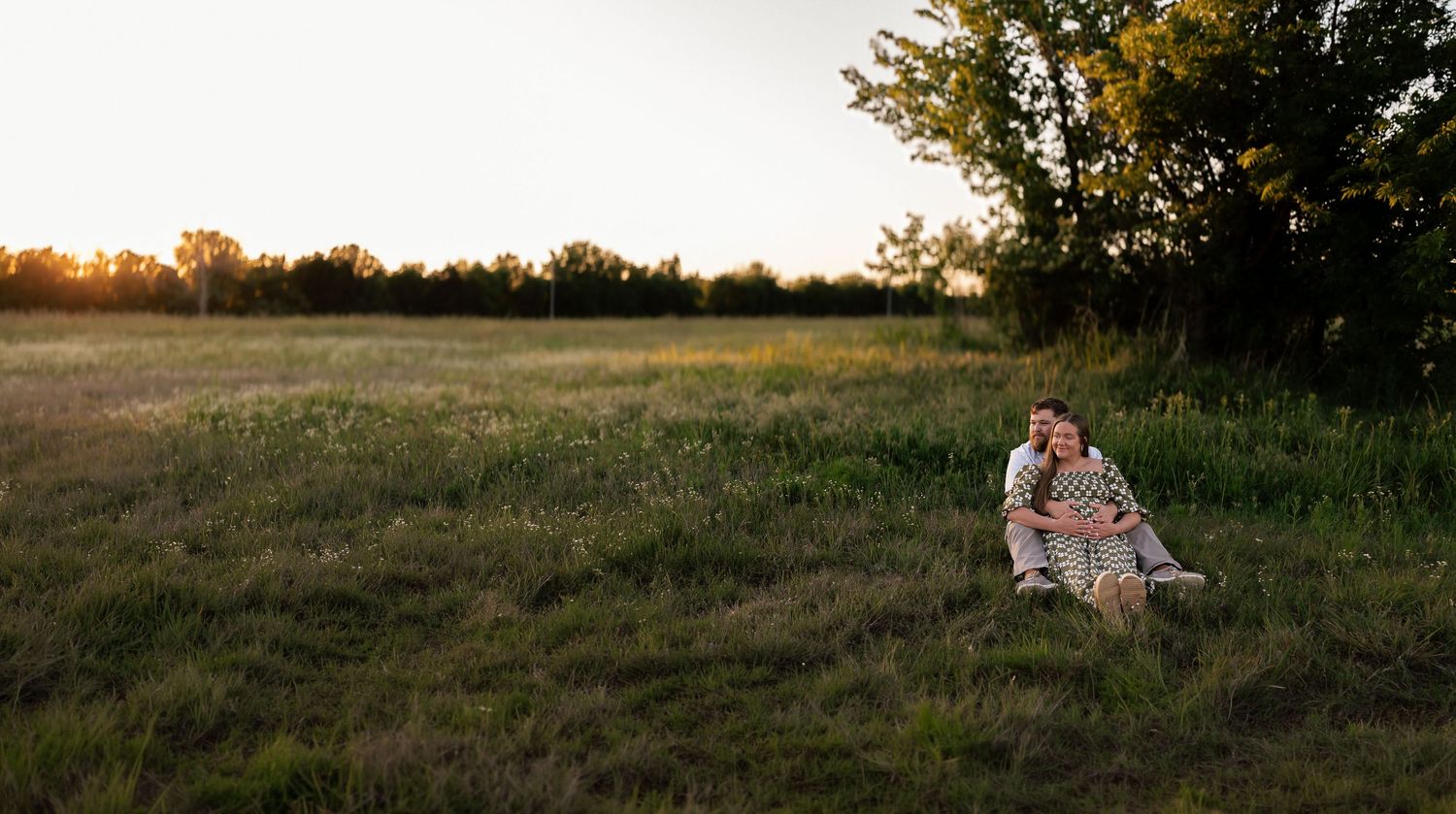 Couple sitting together on a grassy field at golden sunset, surrounded by lush green trees in a romantic outdoor setting.