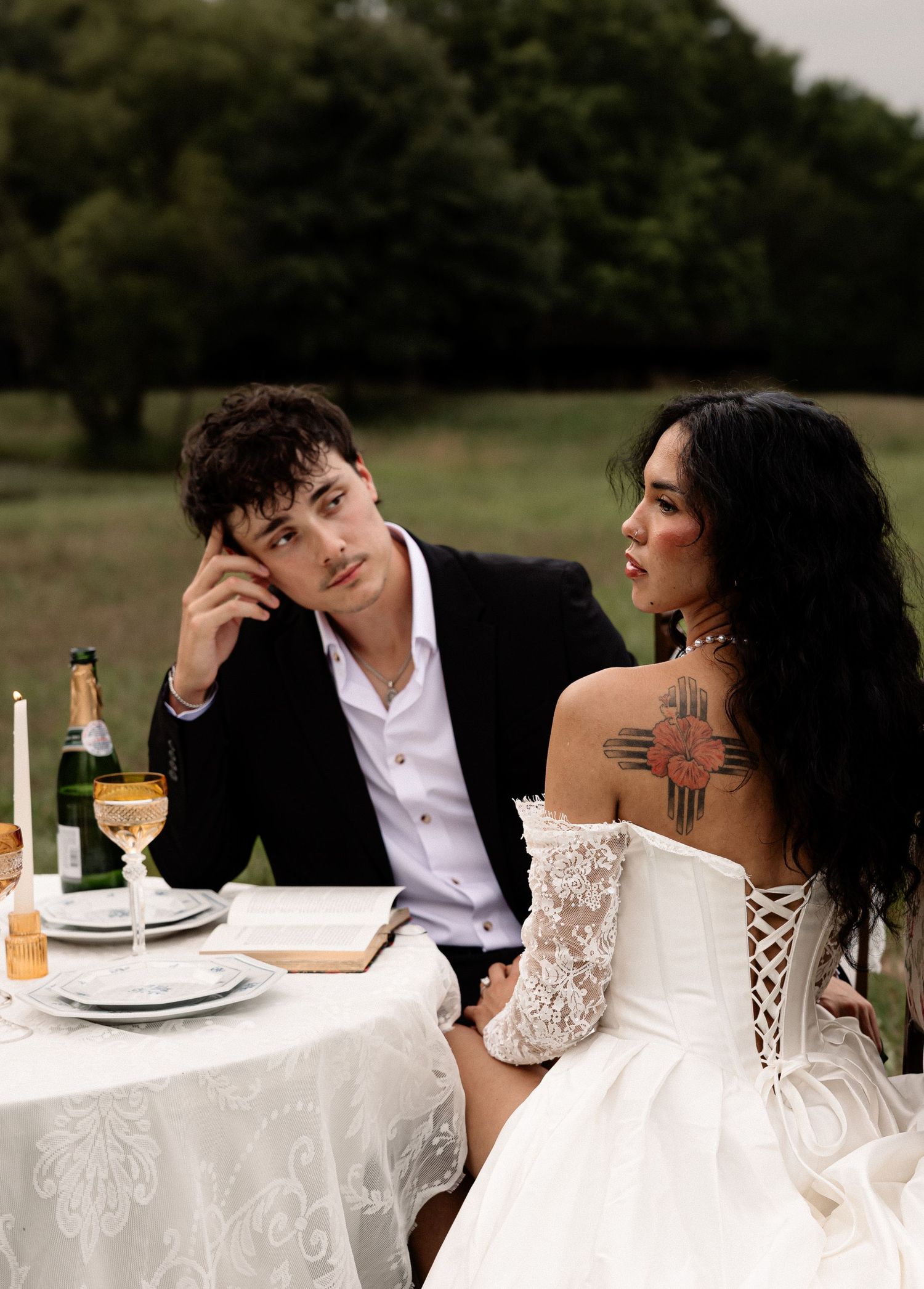 A couple in wedding attire sits at an outdoor table with champagne, the groom in a suit, bride in a lace off-shoulder gown.