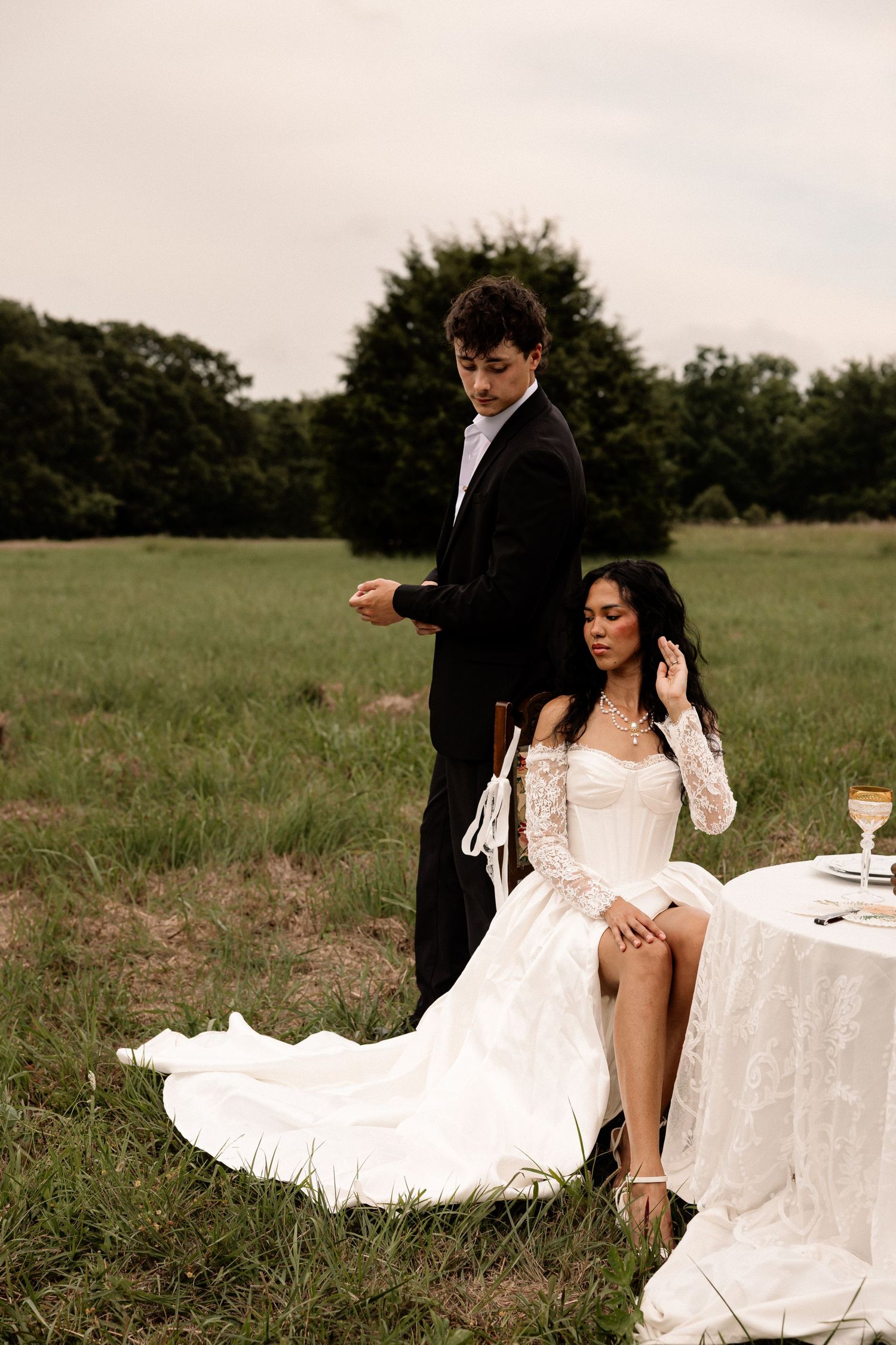 A bride in a white lace gown sits outdoors as her groom in a black suit stands behind her in a green field.