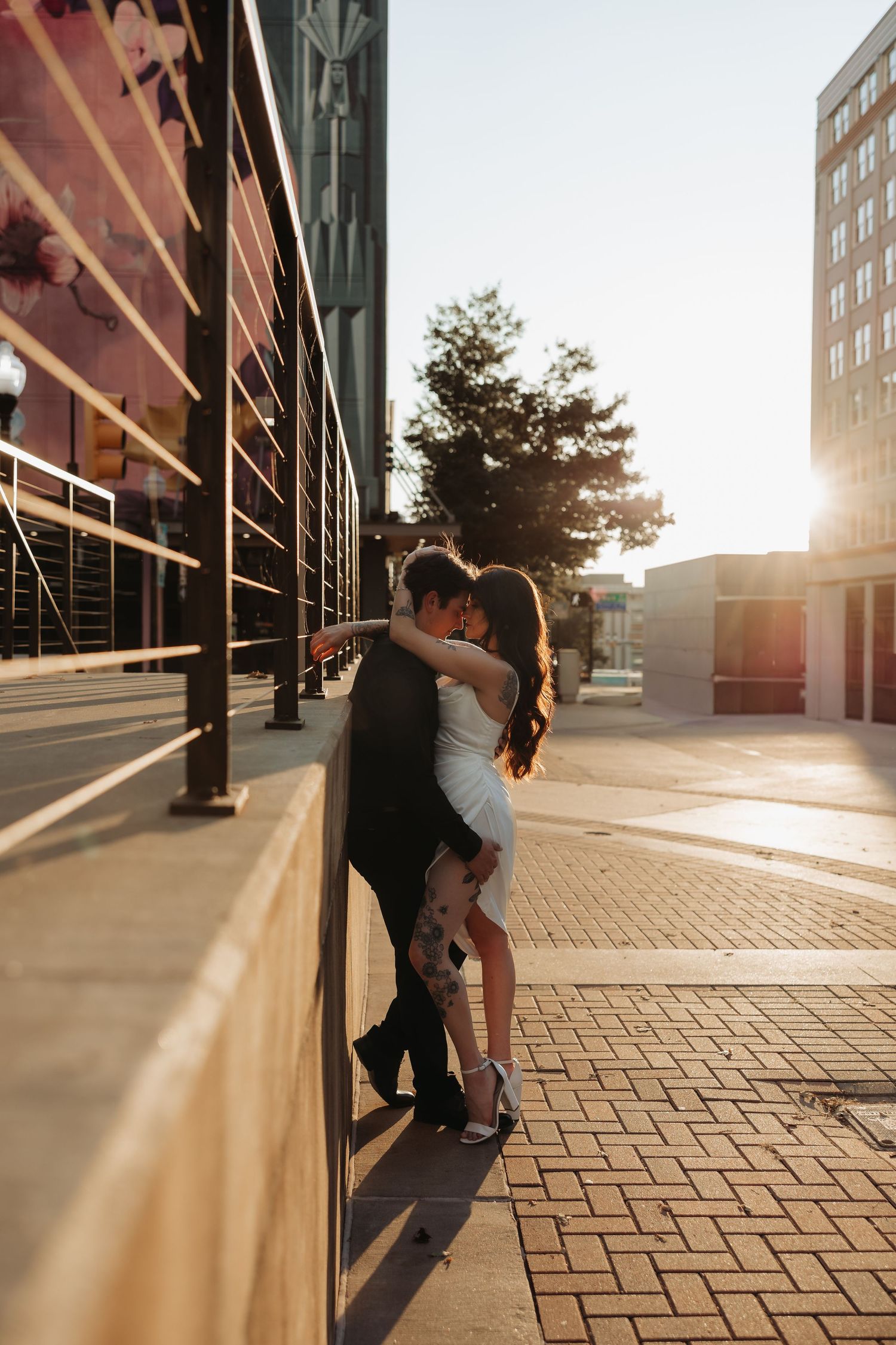 Couple kissing against urban building wall with warm sunlight glowing behind them at sunset. Tulsa Photographer