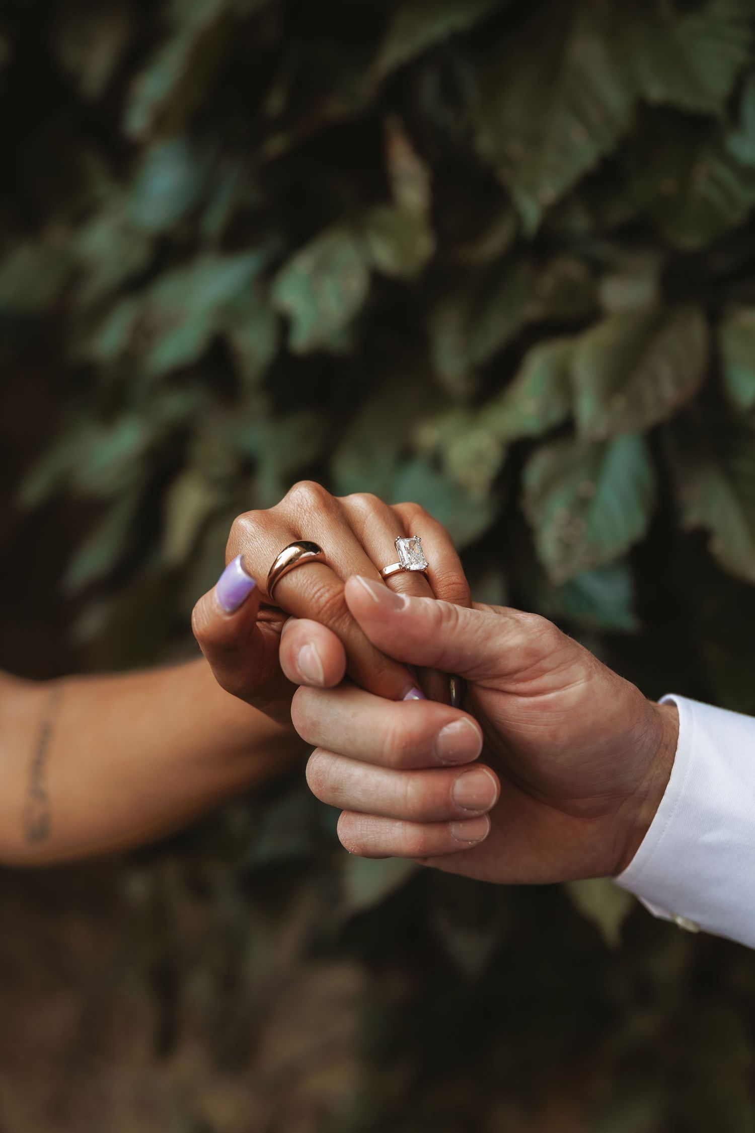 Close-up of newlyweds holding hands, showcasing wedding rings and lavender nail polish against green foliage.