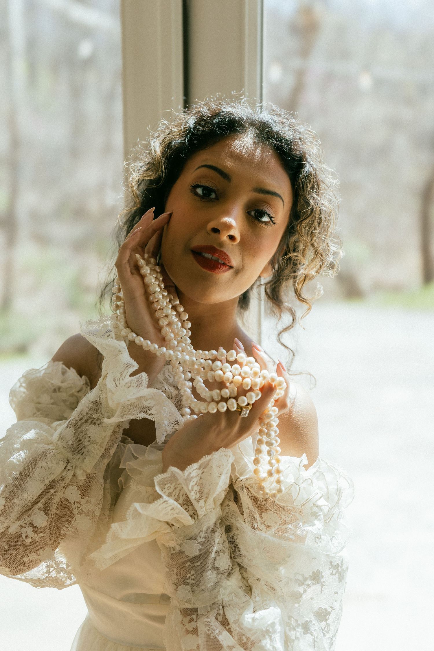 Elegant woman with curly hair wearing white ruffled dress and layered pearl necklaces near a window. Tulsa Photographer