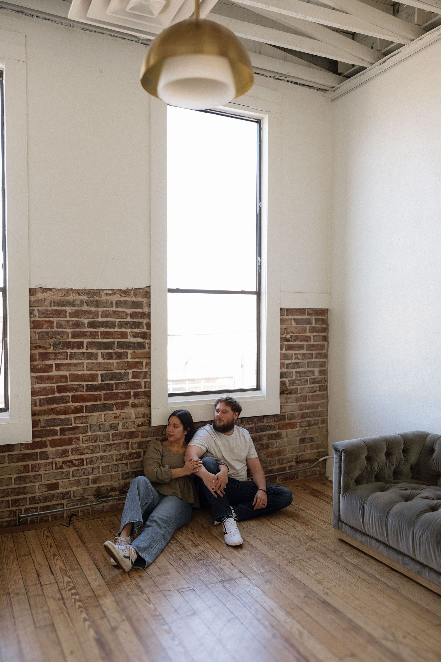 A couple sits together on a hardwood floor in a loft apartment with exposed brick walls and tall windows. Tulsa Photographer