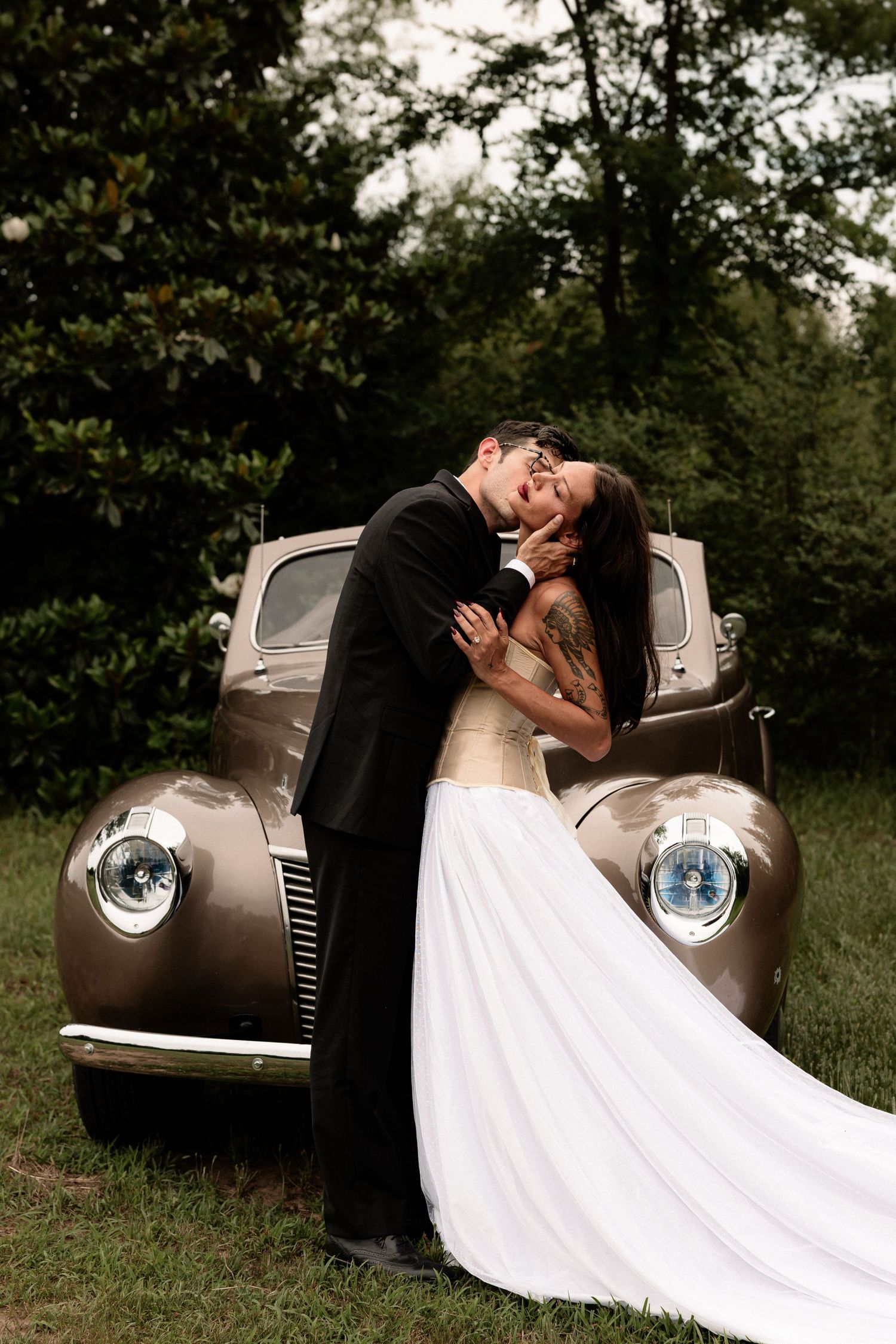 Bride and groom share a romantic kiss leaning against a vintage bronze classic car outdoors surrounded by lush green trees.