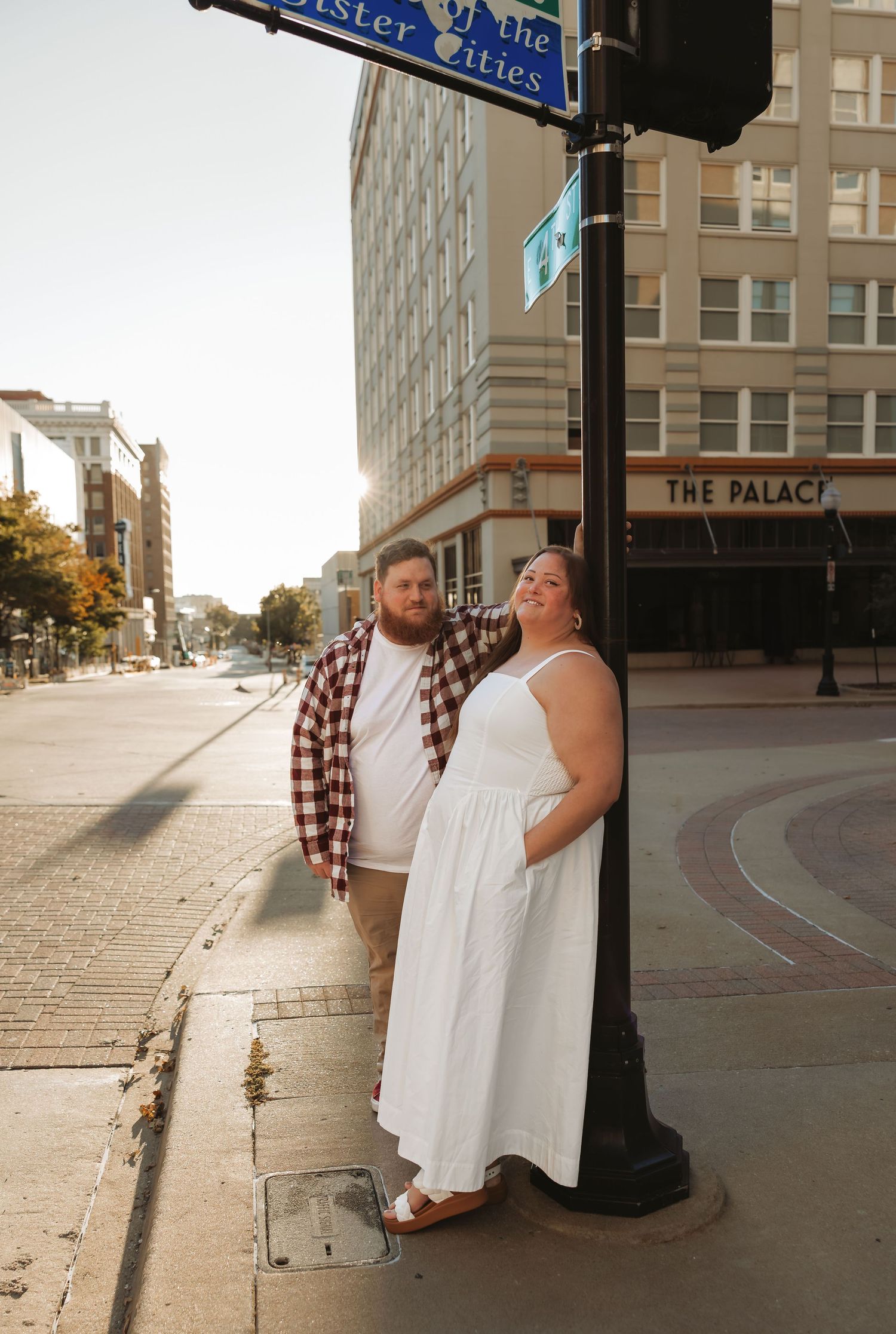 A couple poses together on a sunny urban sidewalk, the woman in white dress leaning against a street pole. Tulsa Photographer