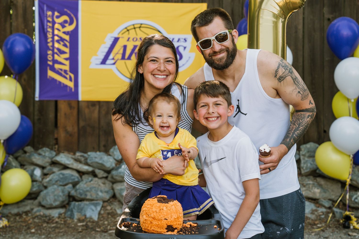 Family of four smiling at a Lakers-themed party with purple and gold balloons and a Lakers banner in the background.