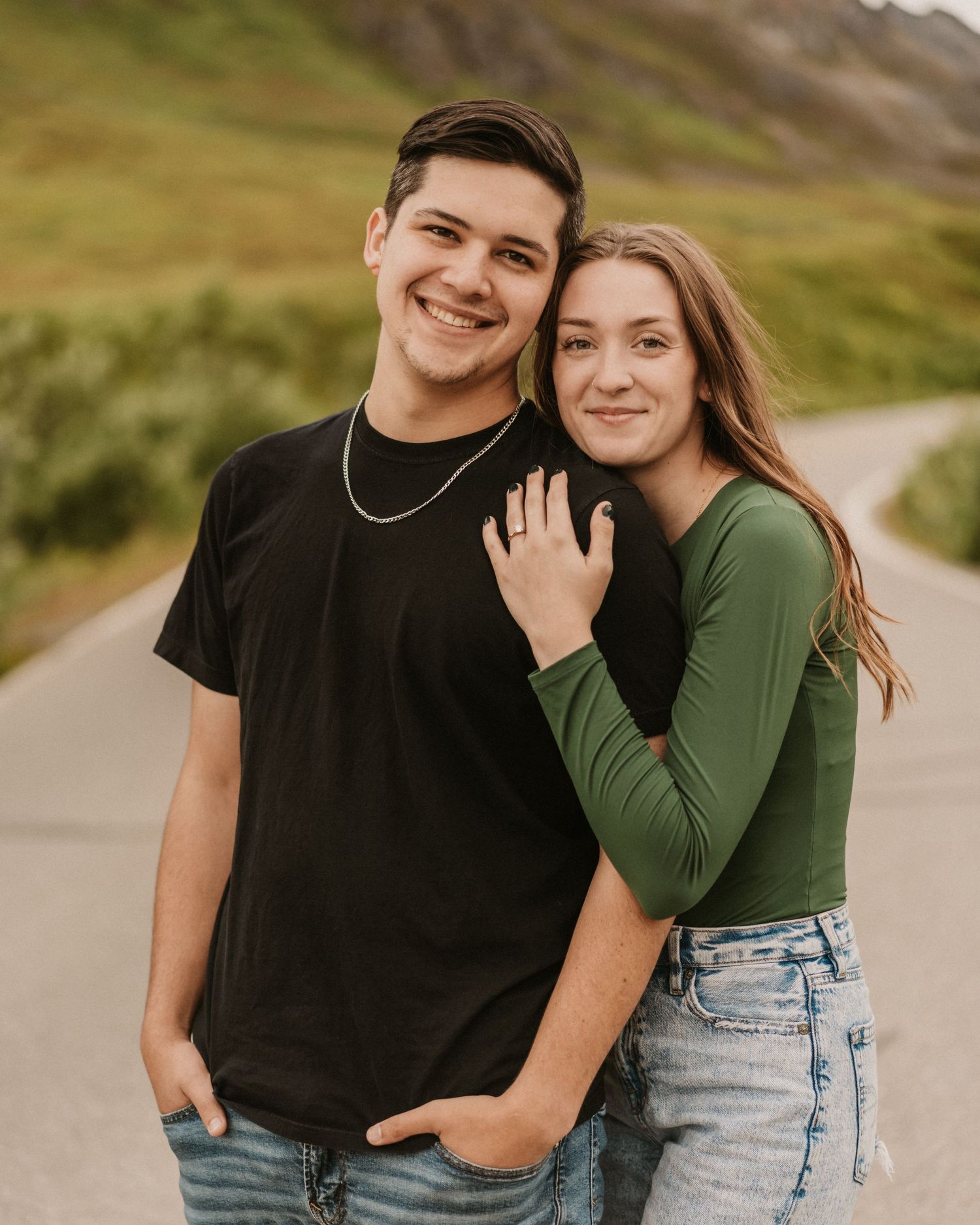Smiling couple posing together on a mountain road surrounded by green hills. Anchorage-Couple-Photographer