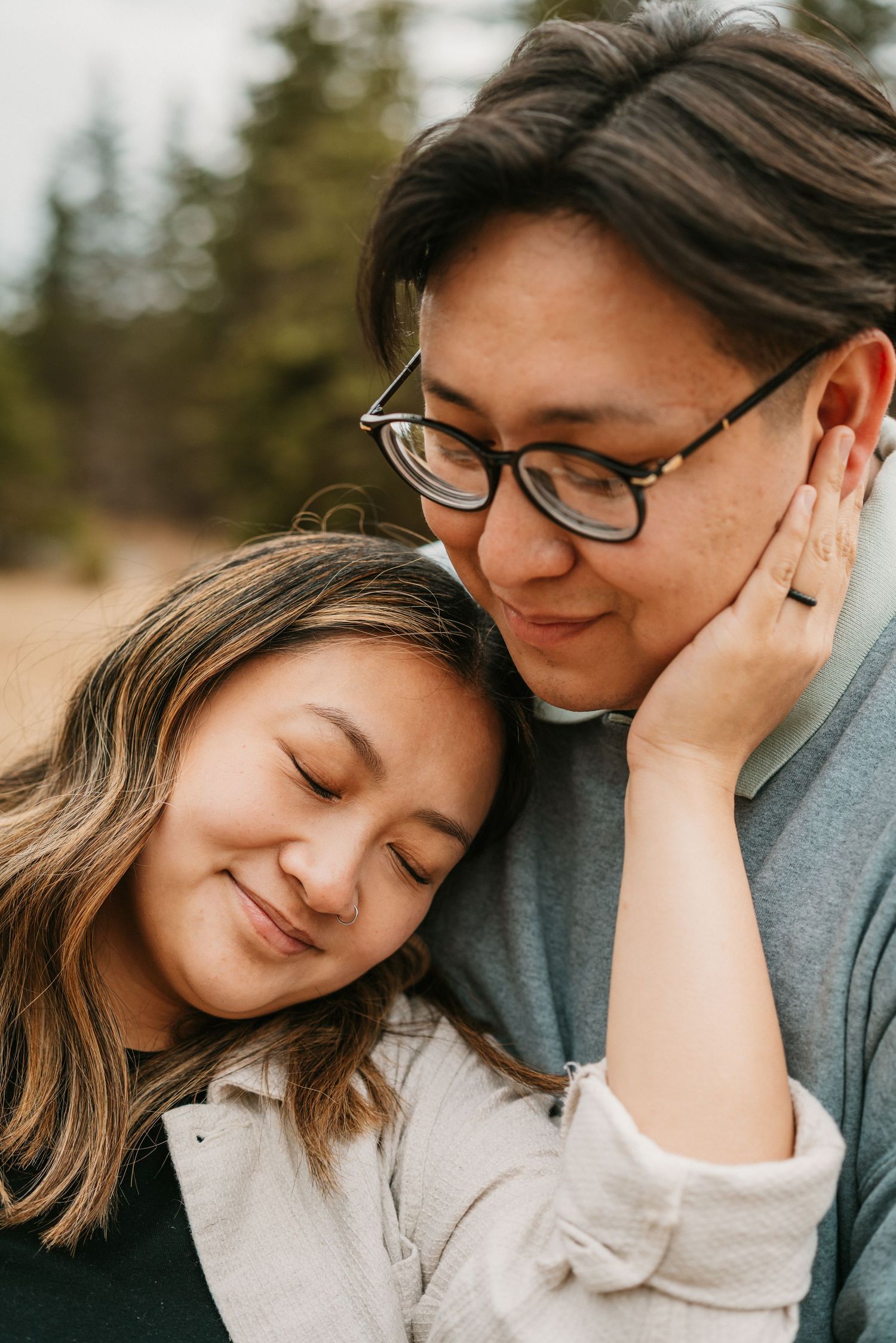 A couple shares a tender moment outdoors, woman resting her head on man's shoulder, both smiling softly.