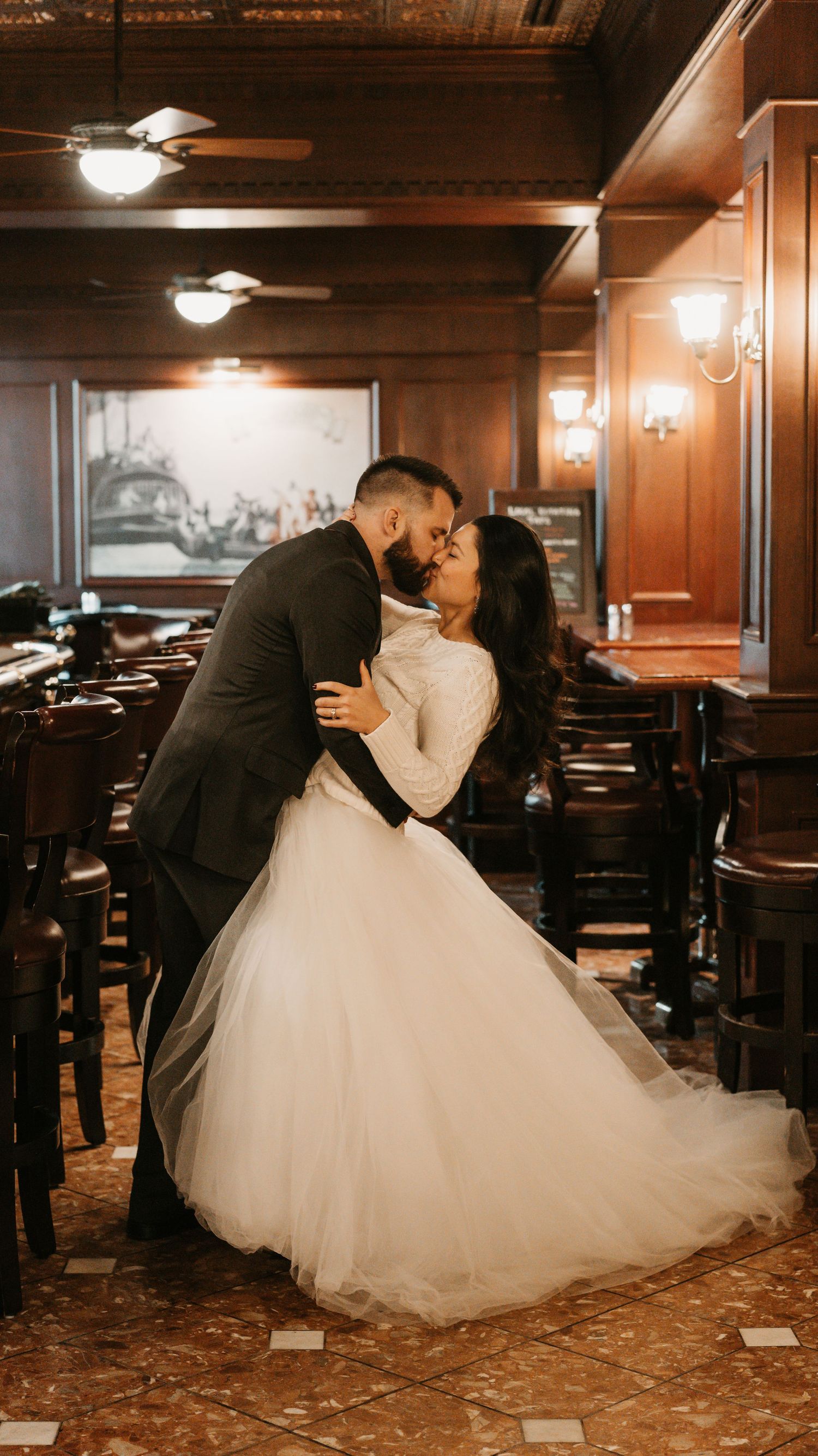 Bride and groom share a romantic kiss in an elegant wood-paneled restaurant, bride wearing a flowing white tulle ballgown.