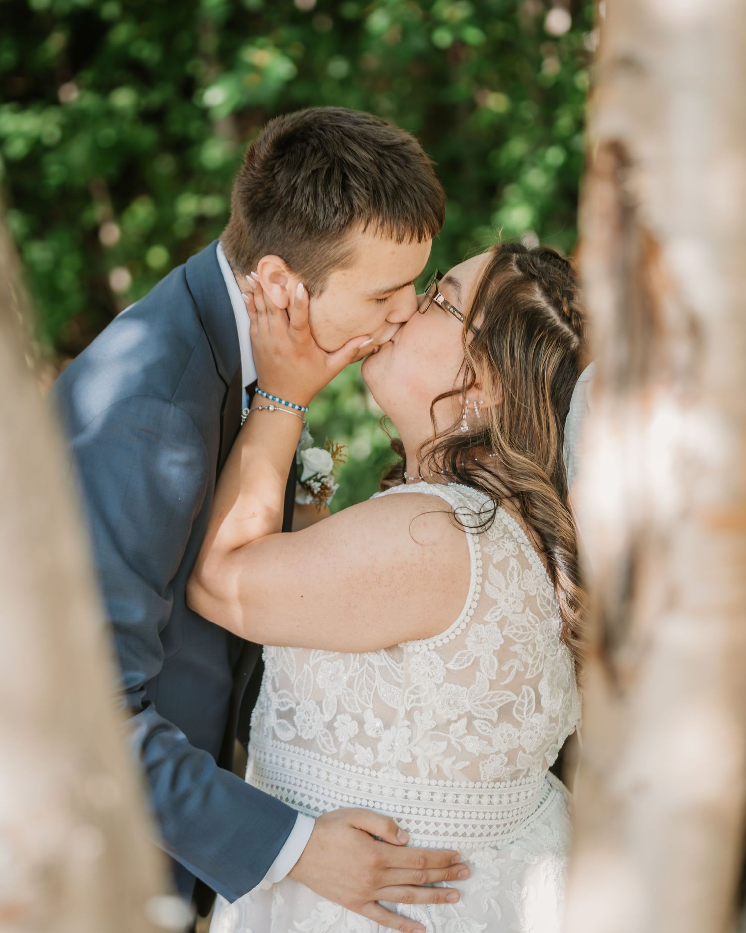 A bride and groom share a romantic kiss outdoors, framed by birch trees, with lush green foliage in the background.