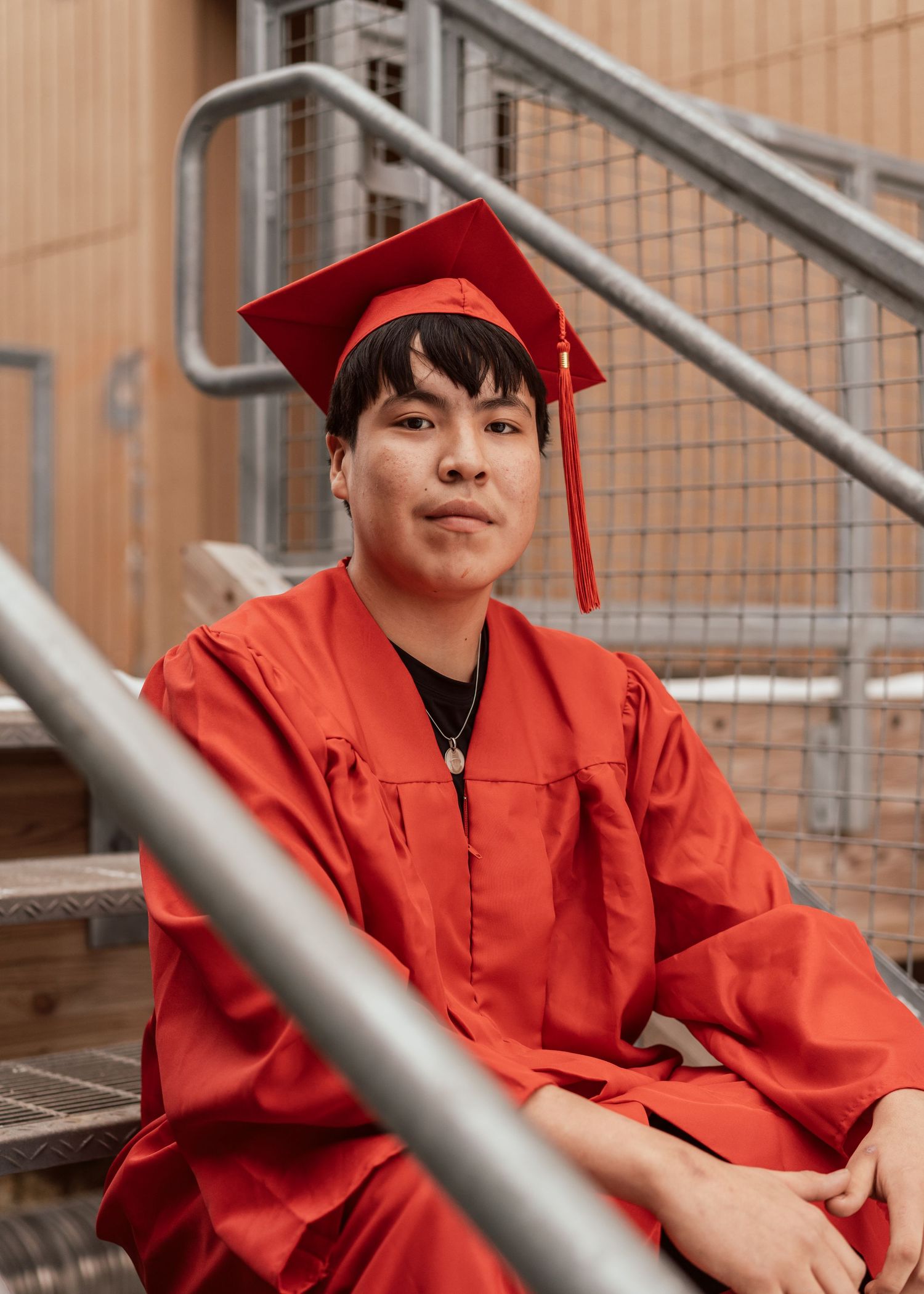Young graduate in red cap and gown sitting on metal stairs, smiling confidently at camera.